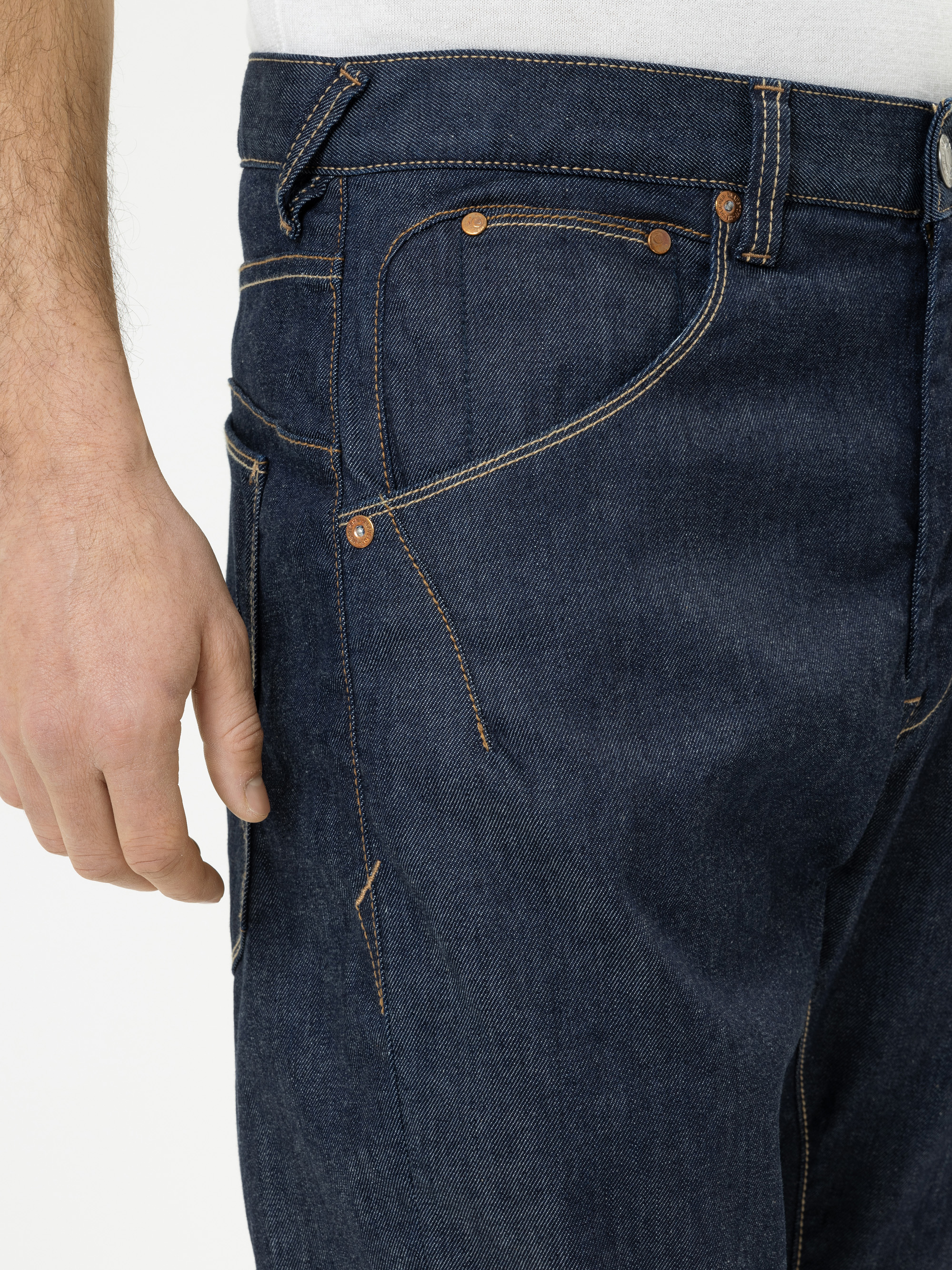 Nahaufnahme einer Person, die eine dunkelblaue Jeans und ein weißes Hemd trägt, wobei die seitlichen und vorderen Taschen mit orangefarbenen Nähten zu sehen sind. Der linke Arm der Person ist neben der Jeans zu sehen.
