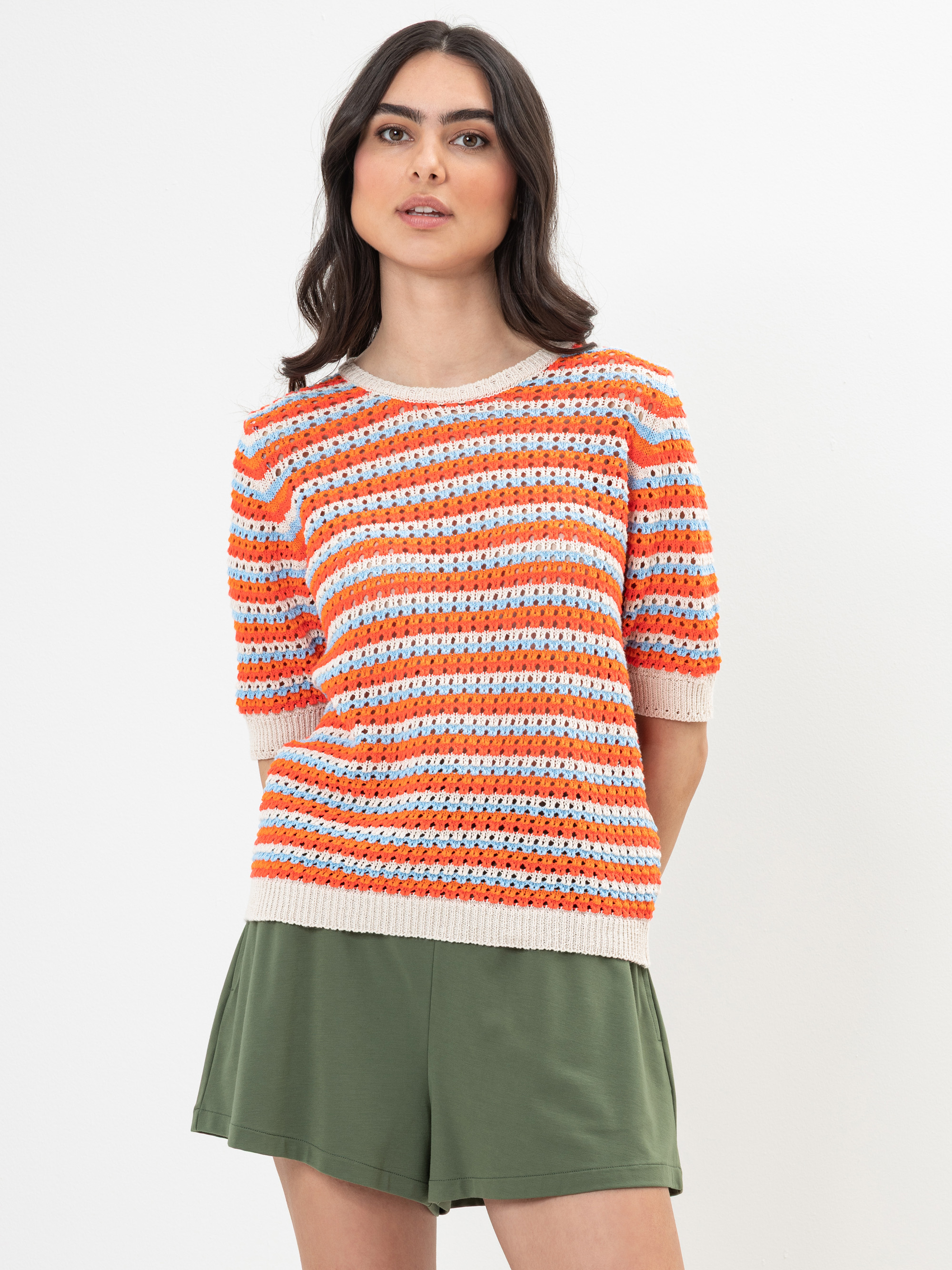 Eine Frau mit gewelltem dunklem Haar trägt ein kurzärmeliges, orange, weiß und blau gestreiftes Strickoberteil und olivgrüne Shorts vor einem schlichten weißen Hintergrund.