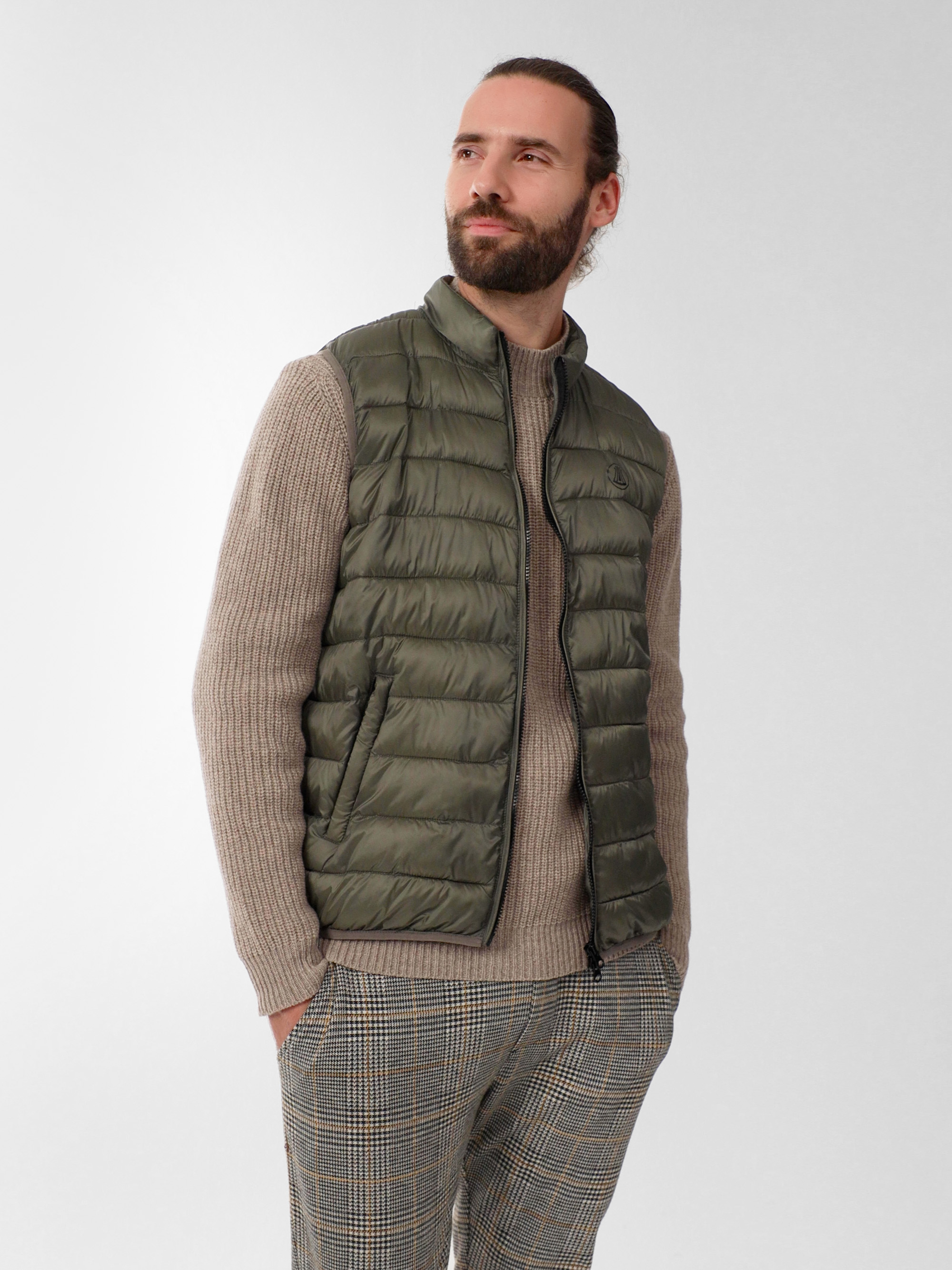 Ein Mann mit Bart und langen, nach hinten gebundenen Haaren trägt eine grüne Pufferweste über einem beigen Strickpullover und eine karierte Hose. Er hat die Hände in den Taschen und blickt leicht zur Seite, während er vor einem schlichten Hintergrund