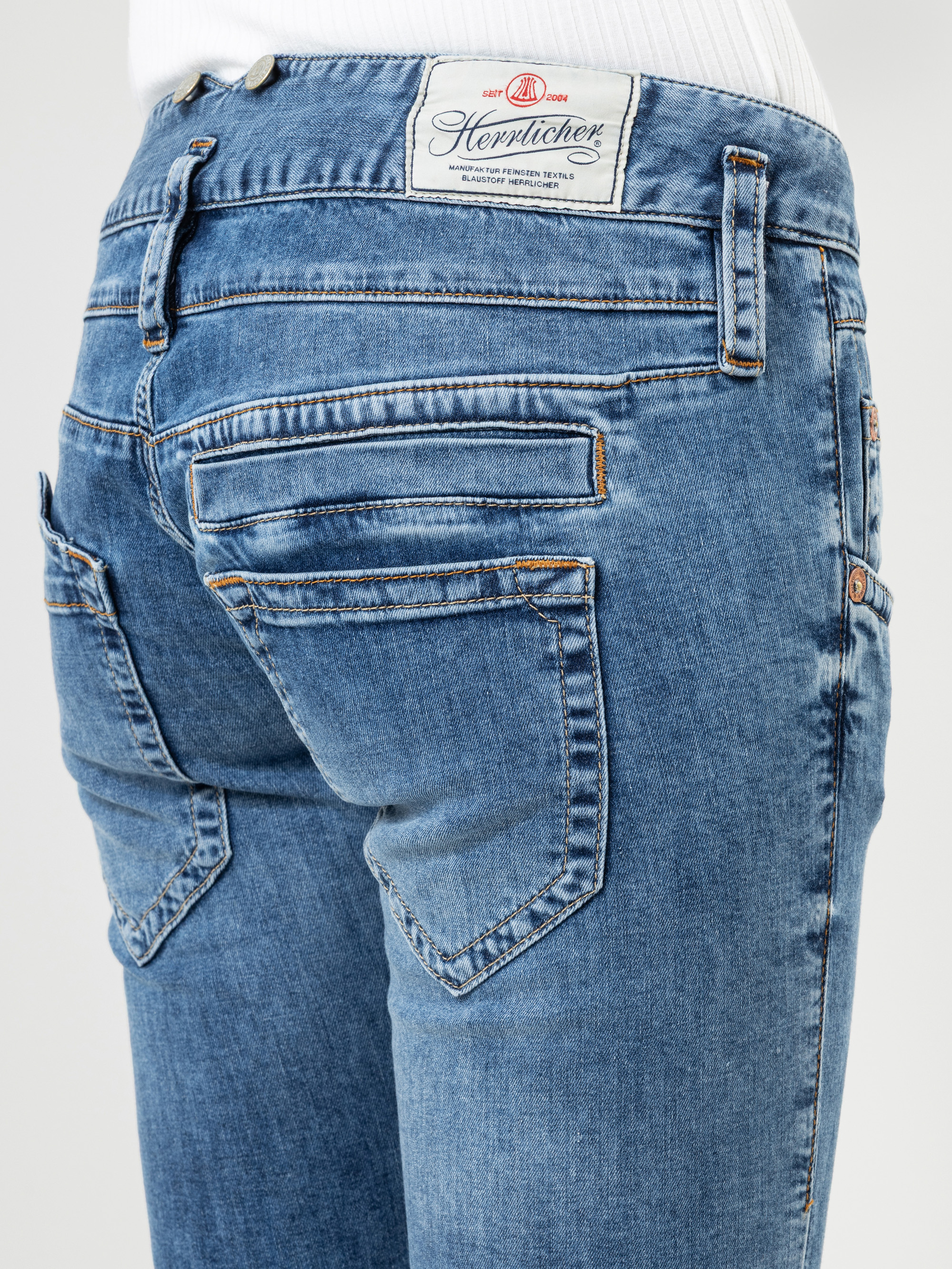 Nahaufnahme des Rückens einer Person, die eine blaue Denim-Jeans mit mehreren Taschen und einem weißen Herrlicher-Markenetikett am Bund trägt. Die Person trägt außerdem ein weißes Oberteil.