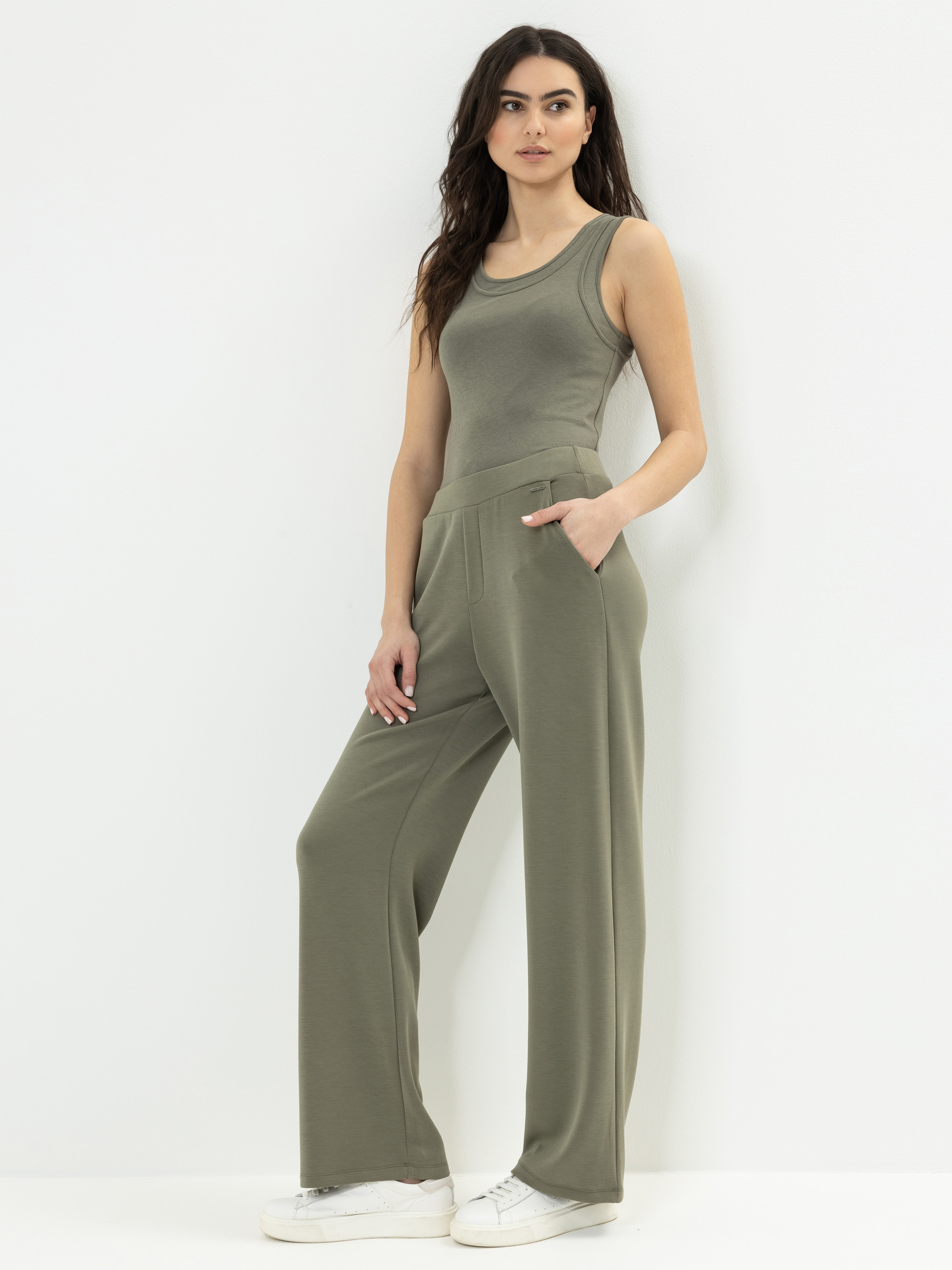 Eine Frau steht vor einer weißen Wand und trägt ein olivgrünes, ärmelloses Tank-Top und eine dazu passende Hose mit weiten Beinen und Taschen. Sie hat lange dunkle Haare und trägt weiße Turnschuhe.