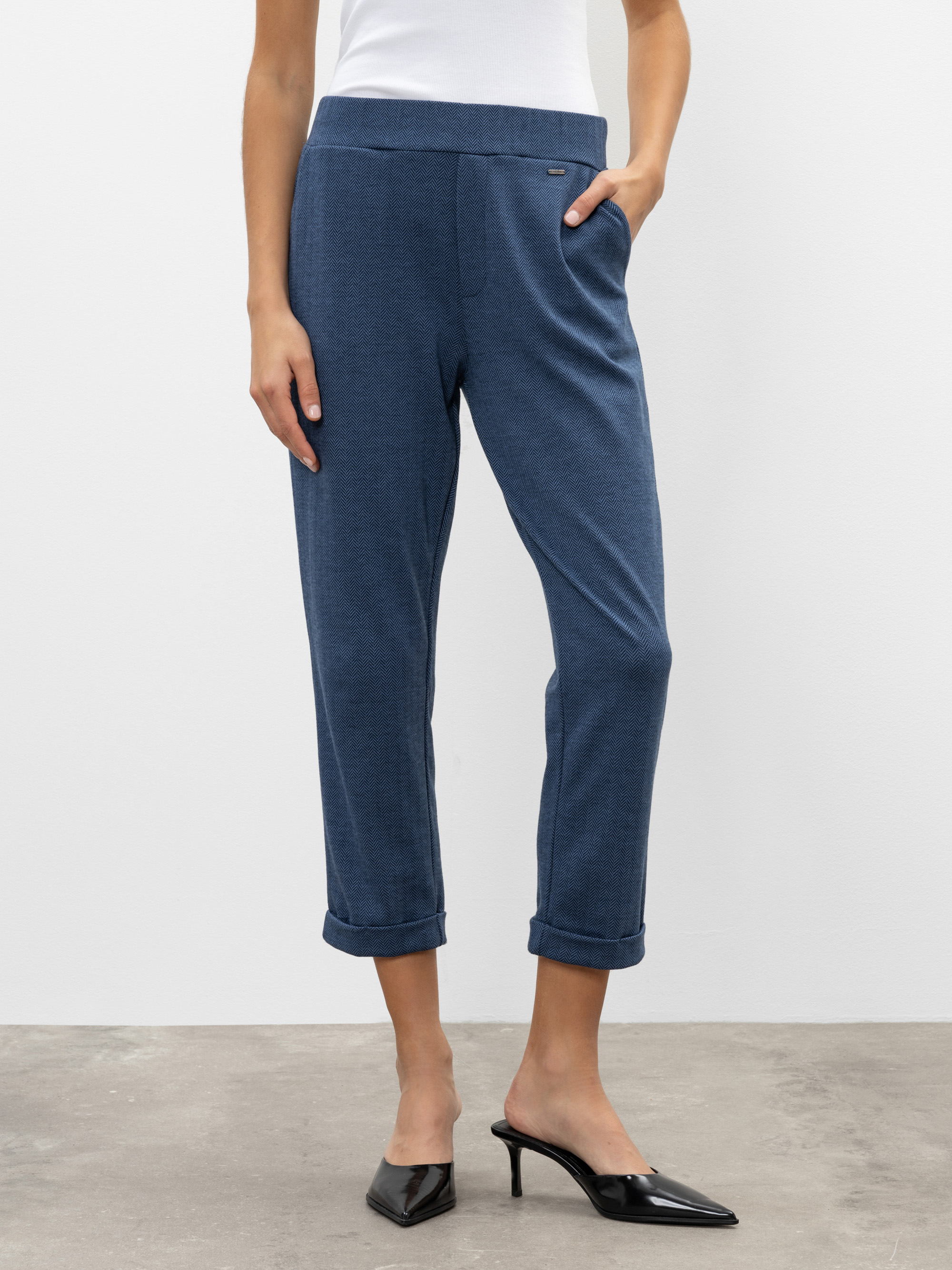 Eine Person, die eine blaue knöchellange Hose mit aufgerollten Manschetten, ein weißes Oberteil und schwarze Pantoletten mit Absätzen trägt, steht vor einem schlichten hellen Hintergrund und hat eine Hand in der Tasche.