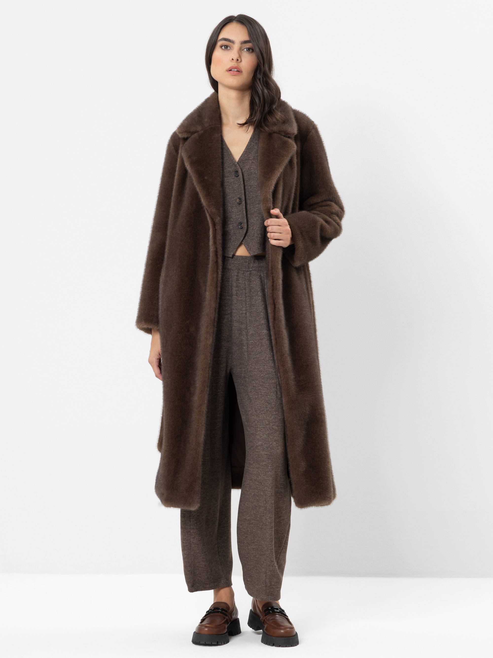 Vor einem weißen Hintergrund steht eine Frau, die einen langen braunen Kunstpelzmantel über einem braunen Strickpullover, einer dazu passenden Hose und braunen Loafern trägt. Sie hat langes dunkles Haar, einen ausdruckslosen Gesichtsausdruck und hält