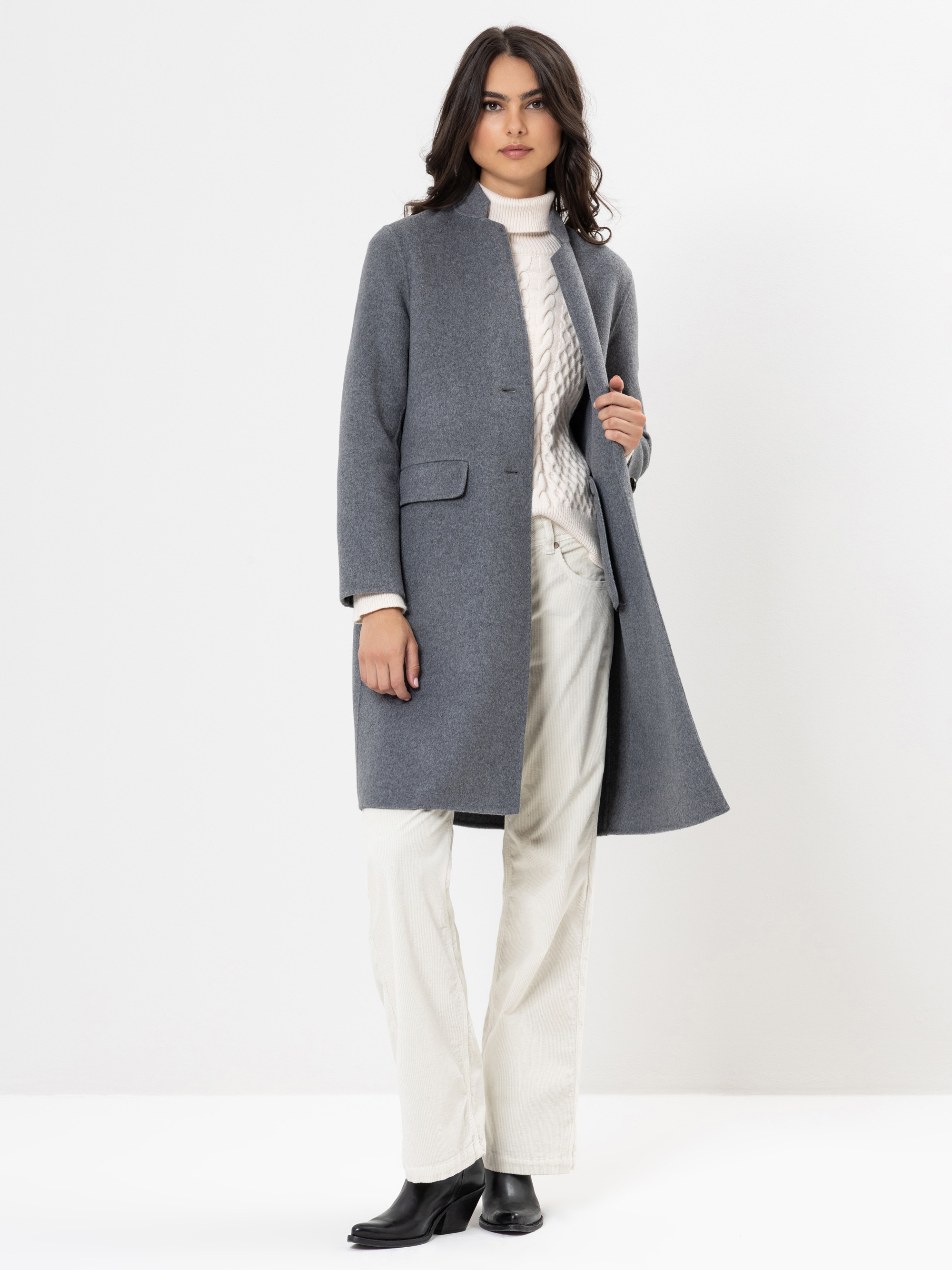Eine Frau, die einen grauen Mantel über einem weißen Rollkragenpullover und eine weiße Hose, gepaart mit schwarzen Stiefeletten, trägt, steht vor einem schlichten weißen Hintergrund.