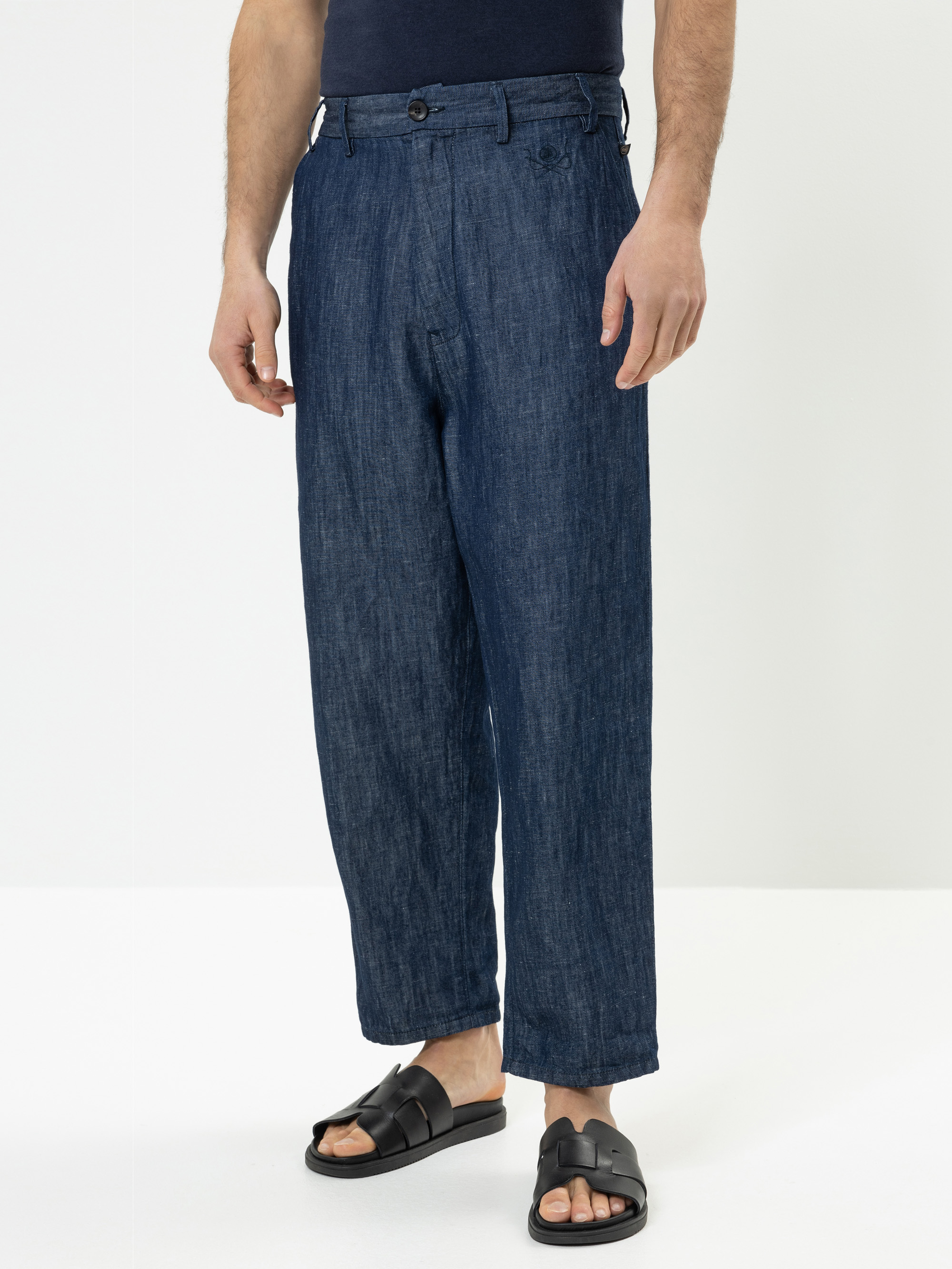 Eine Person, die eine locker sitzende, kurz geschnittene blaue Jeanshose, ein dunkles Hemd und schwarze Gleitsandalen trägt, steht vor einem schlichten weißen Hintergrund. Nur der Unterkörper ist sichtbar.