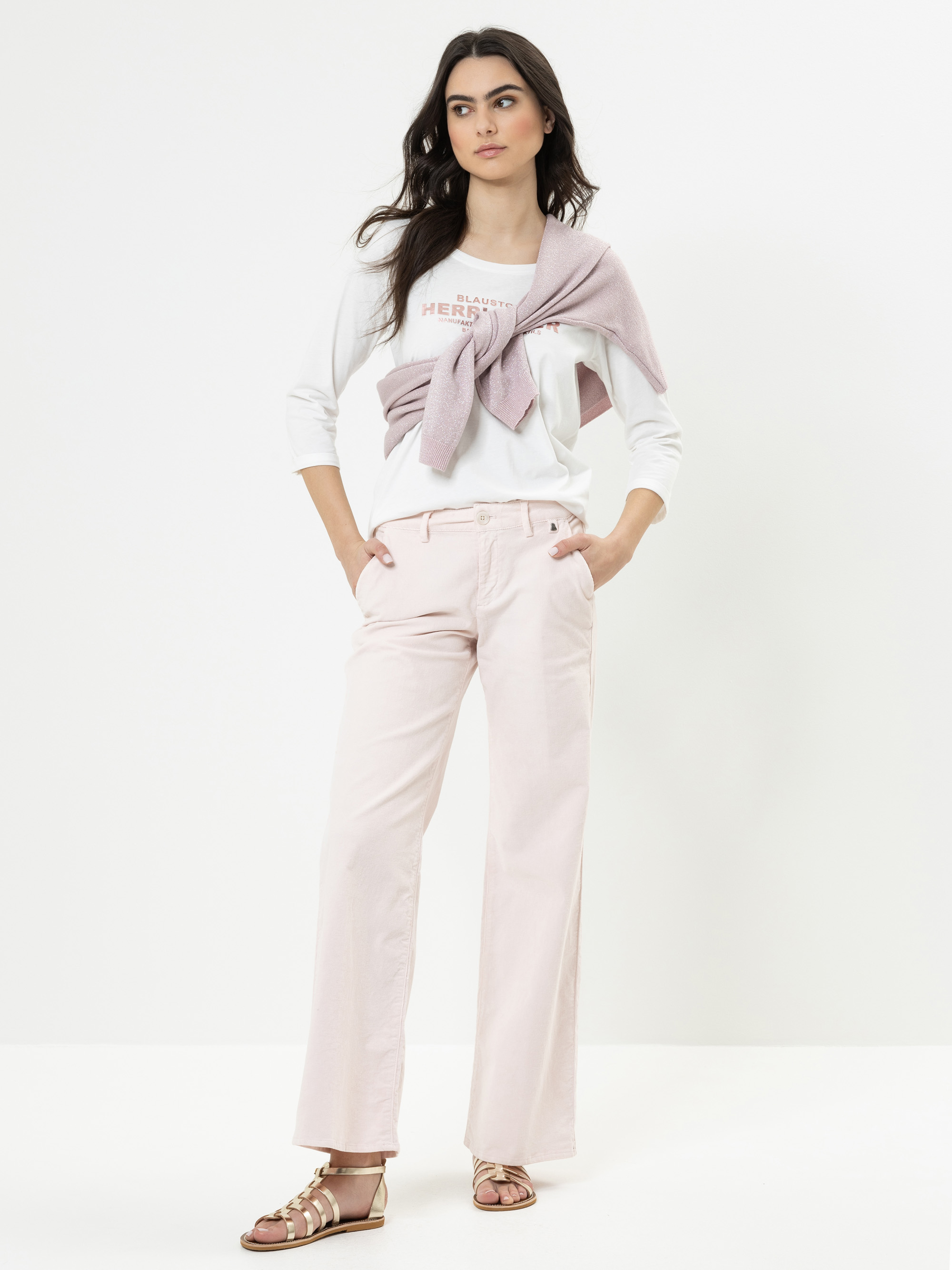 Eine Frau steht vor einem weißen Hintergrund und trägt eine hellrosa Hose, ein weißes Oberteil mit einem über die Schultern drapierten rosa Pullover und metallische Sandalen. Ihre Hände stecken in den Taschen und sie hat einen entspannten Gesichtsaus