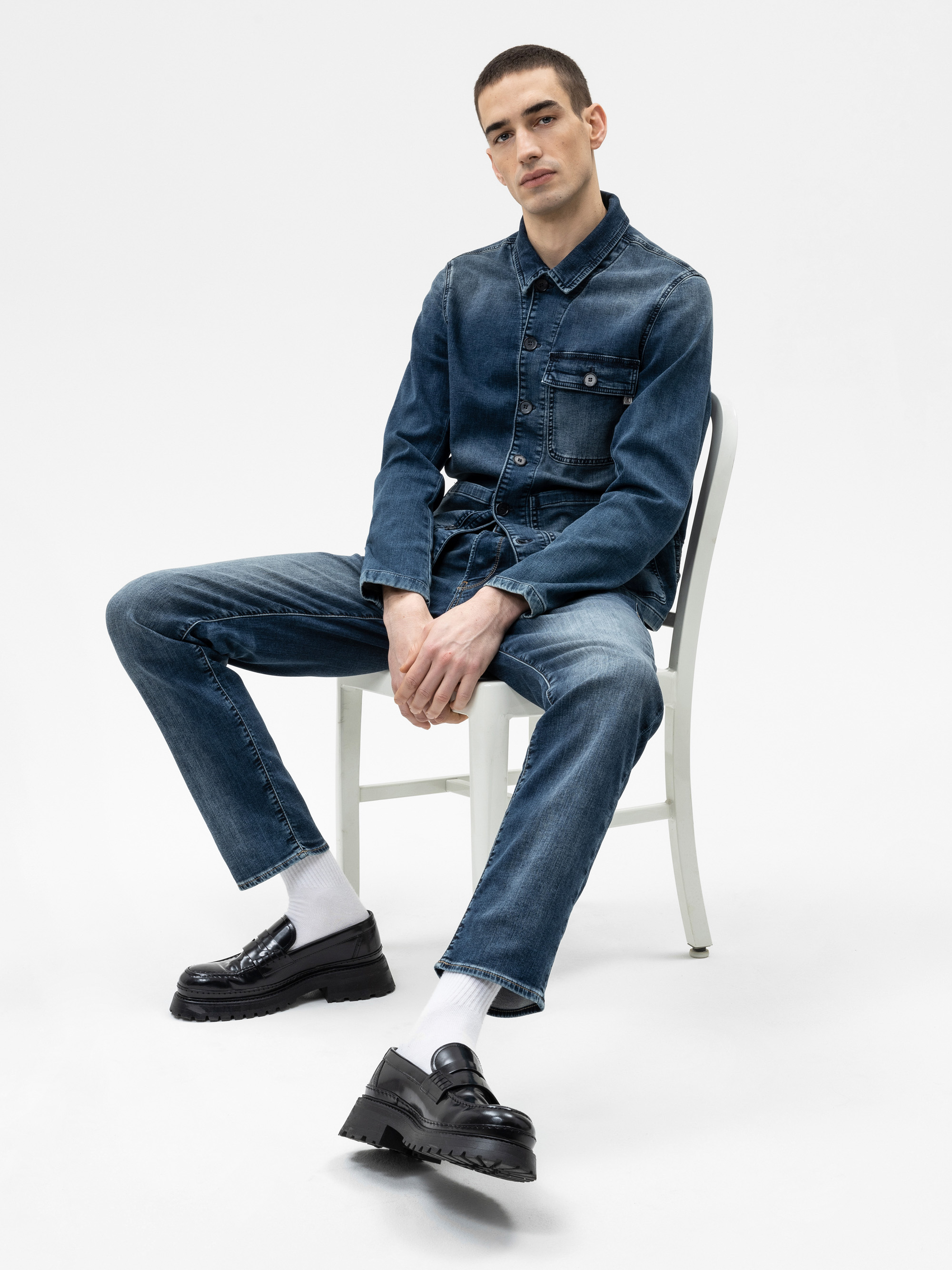 Ein Mann, der ein Jeanshemd und Jeans mit weißen Socken und schwarzen Schuhen trägt, sitzt lässig auf einem weißen Stuhl vor einem schlichten weißen Hintergrund.