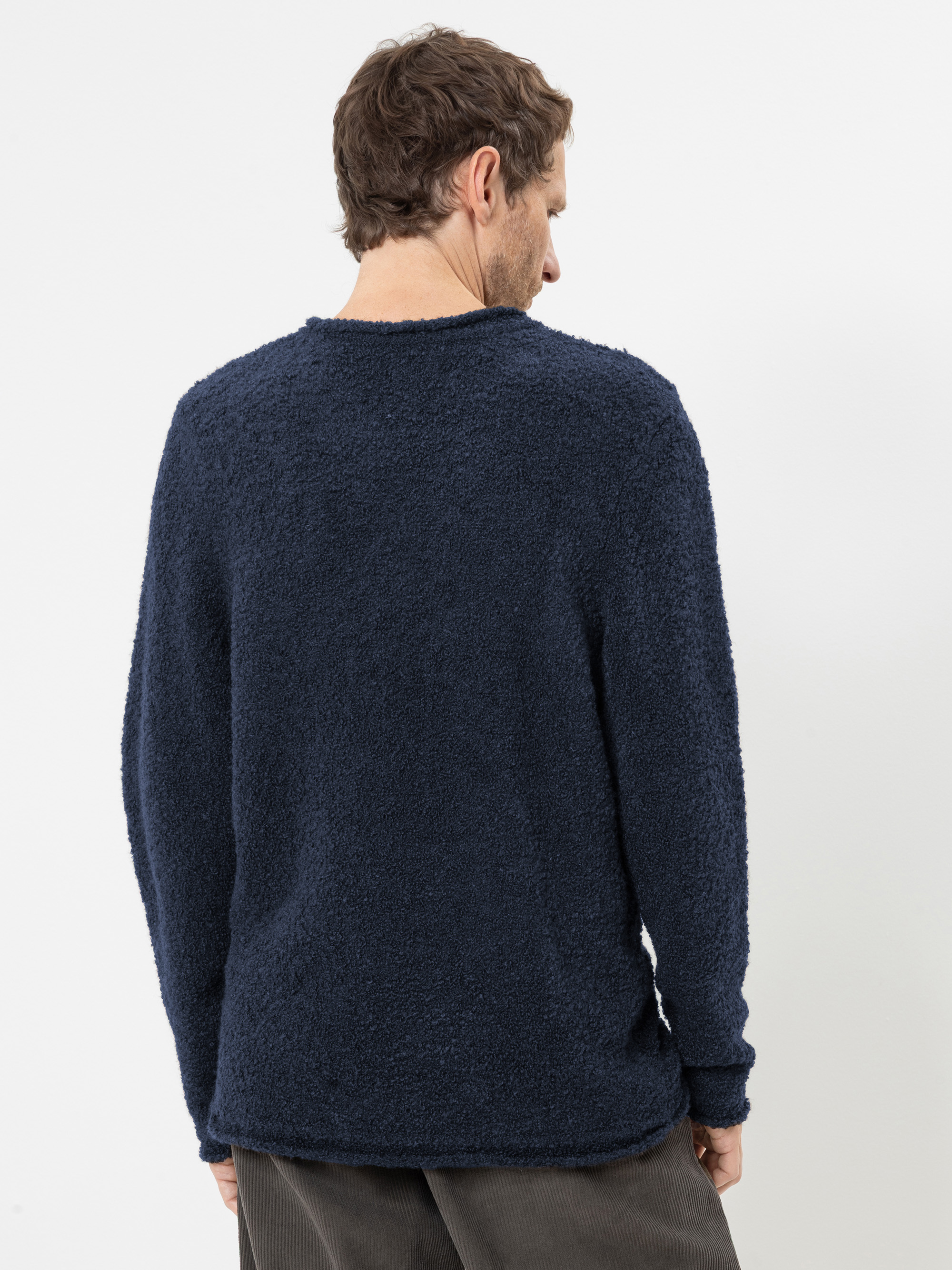 Ein Mann steht mit dem Rücken zur Kamera und trägt einen strukturierten, langärmeligen, marineblauen Pullover sowie eine braune Hose. Der Hintergrund ist schlicht weiß.