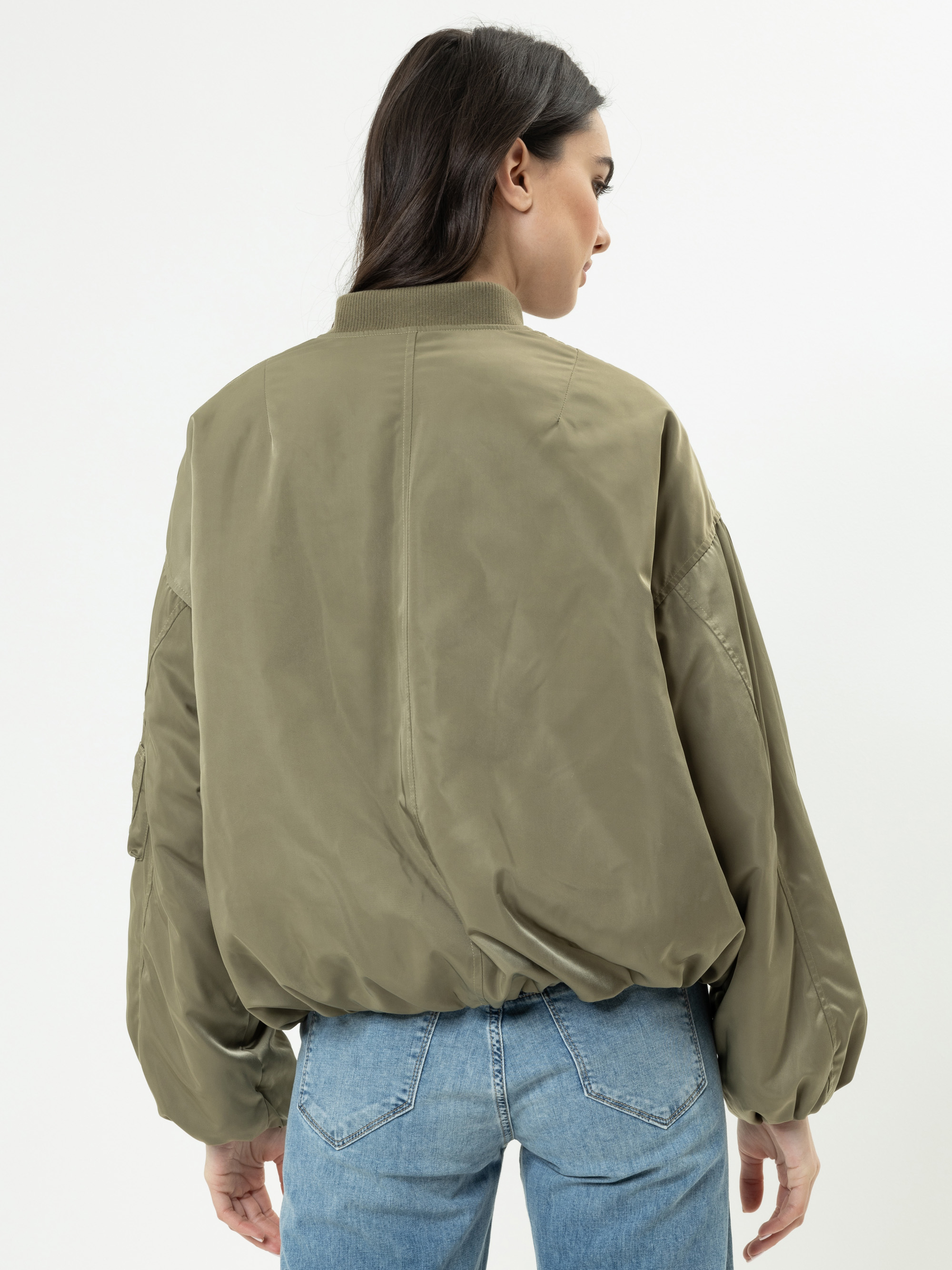 Eine Frau mit langen braunen Haaren trägt eine lockere olivgrüne Bomberjacke und hellblaue Jeans und steht mit dem Rücken zur Kamera vor einem schlichten weißen Hintergrund.