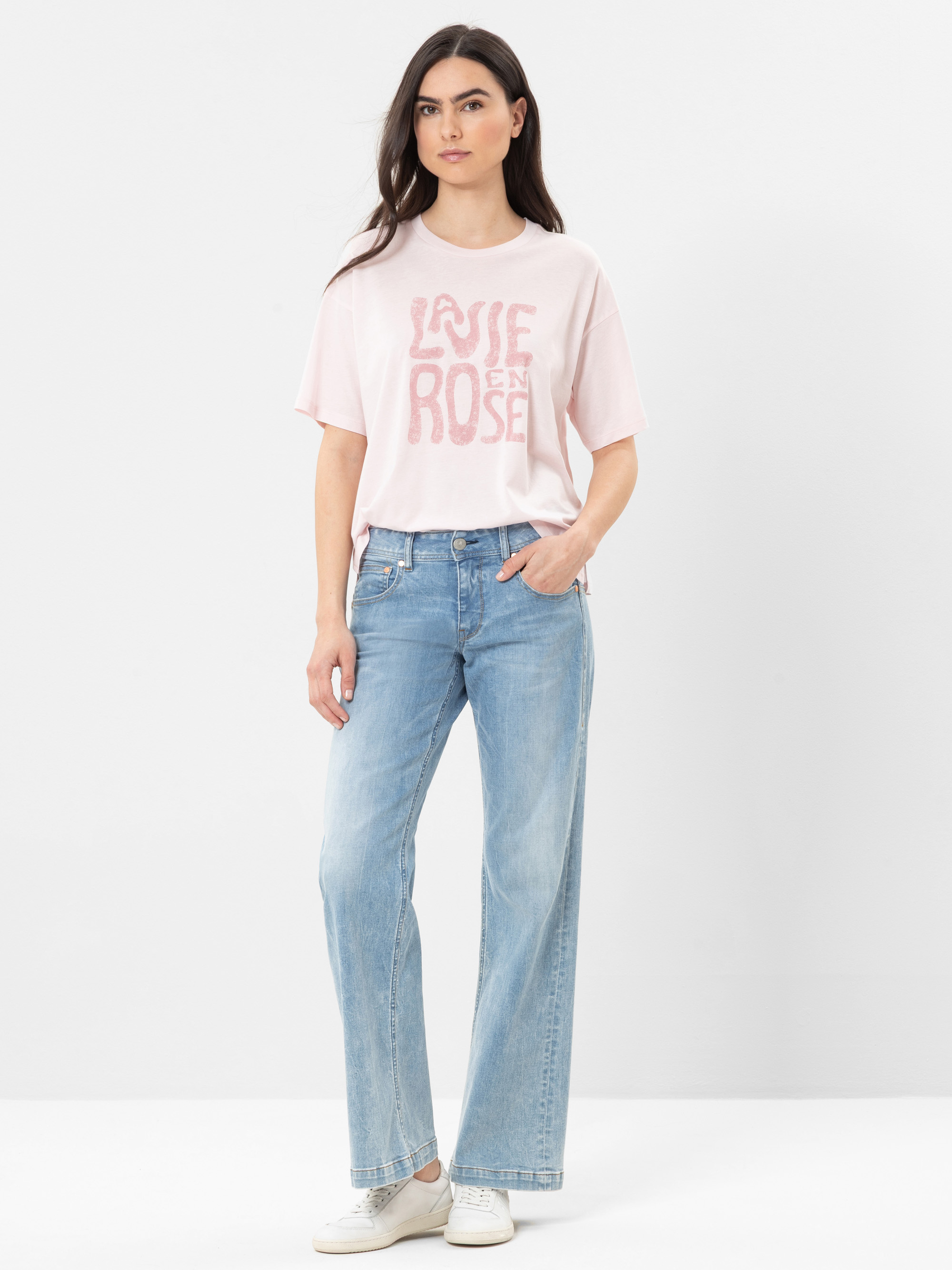 Eine Frau steht vor einem weißen Hintergrund und trägt ein hellrosa T-Shirt mit der Aufschrift La Vie En Rose in fetten Buchstaben, hellblaue Jeans mit weitem Bein und weiße Turnschuhe. Sie hat langes dunkles Haar und einen neutralen Gesichtsausdruck.