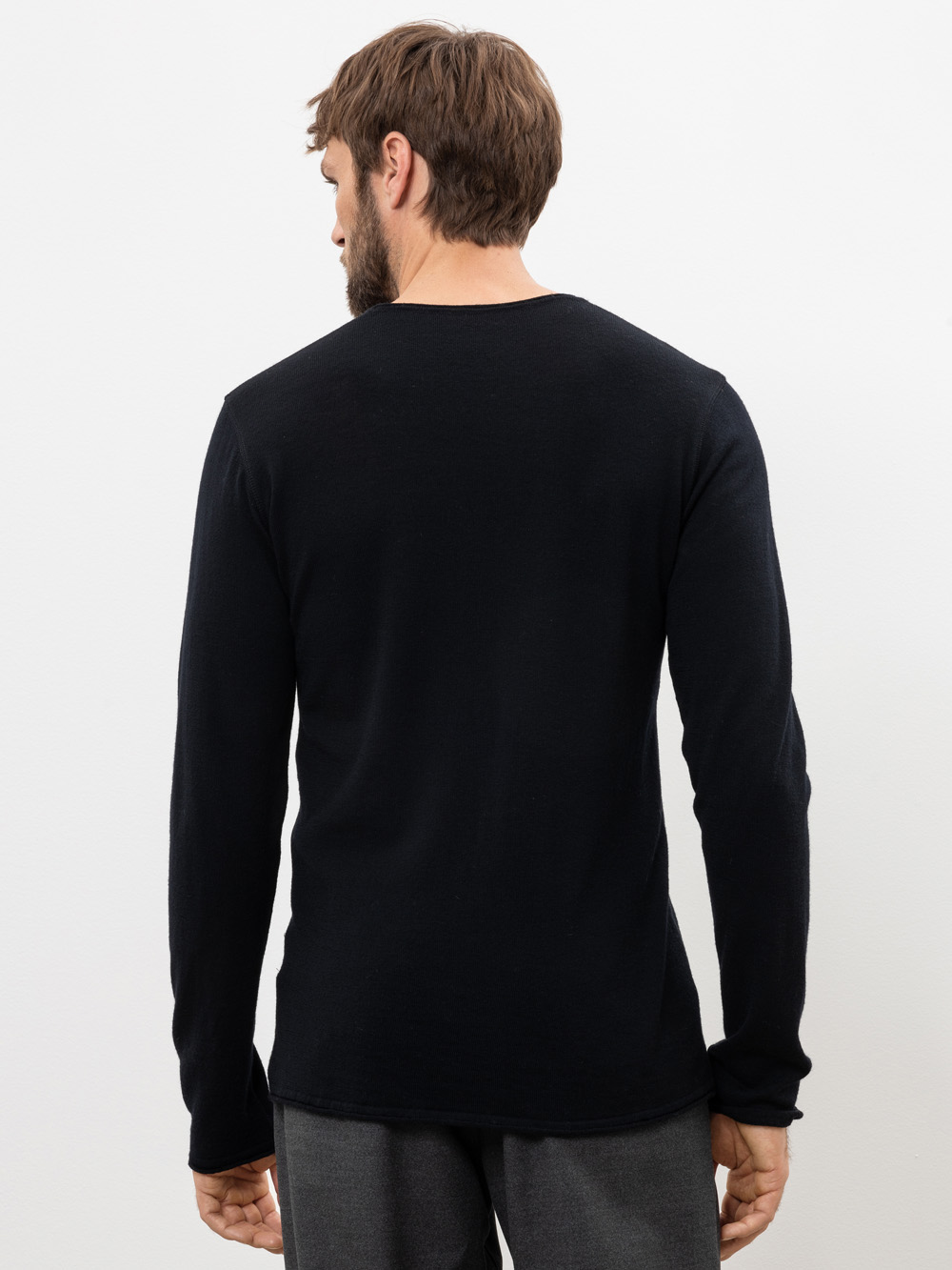 Ein Mann mit kurzen braunen Haaren und Bart steht mit dem Rücken zur Kamera, trägt ein langärmeliges schwarzes Hemd und eine graue Hose vor einem schlichten weißen Hintergrund.