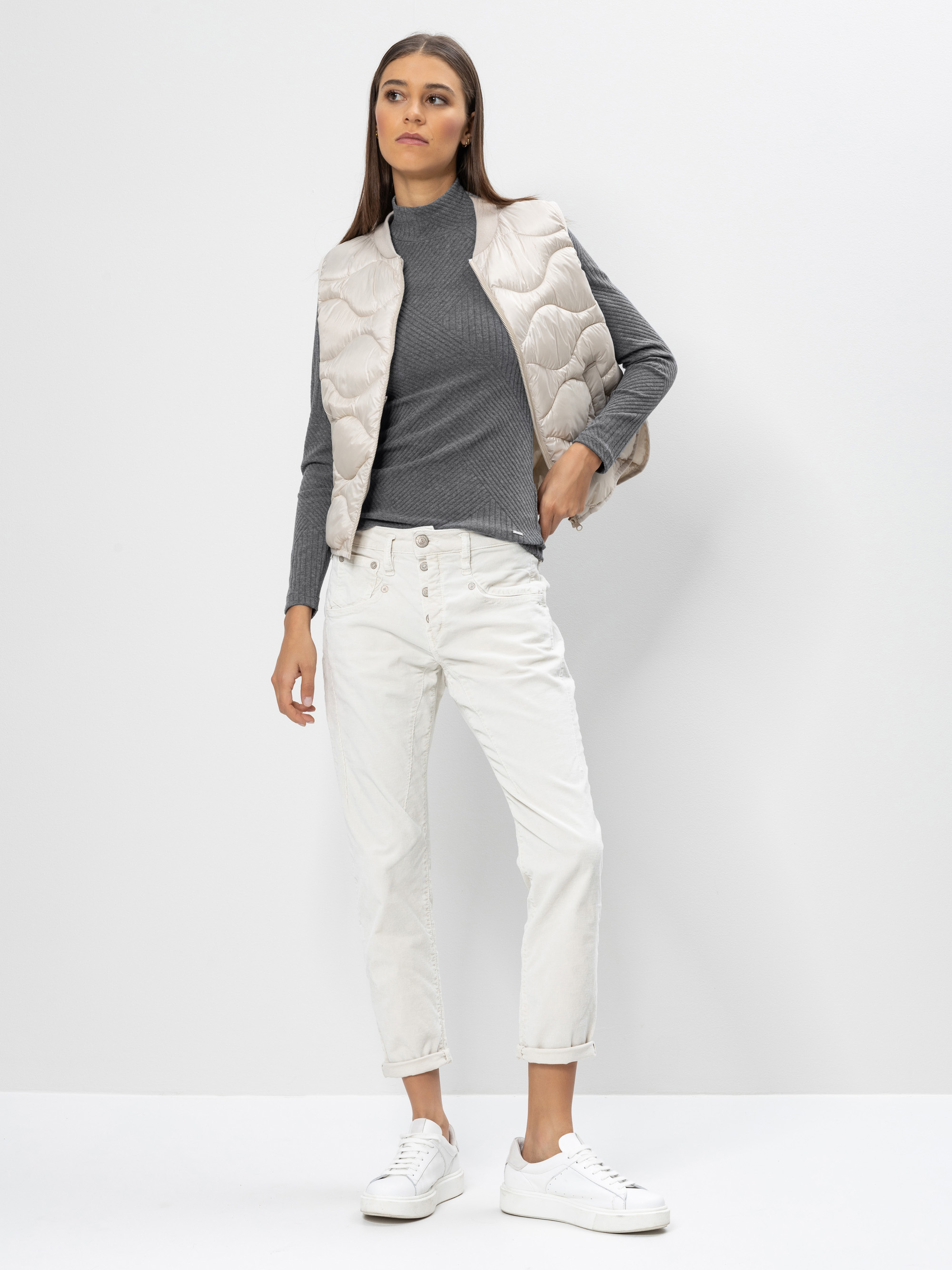 Eine Frau steht vor einem schlichten Hintergrund und trägt eine hellbeige Steppweste, einen grauen Rollkragenpullover, eine weiße Hose mit Knopfleiste und weiße Turnschuhe. Ihre Hand ruht in ihrer Tasche und sie blickt selbstbewusst nach vorne.