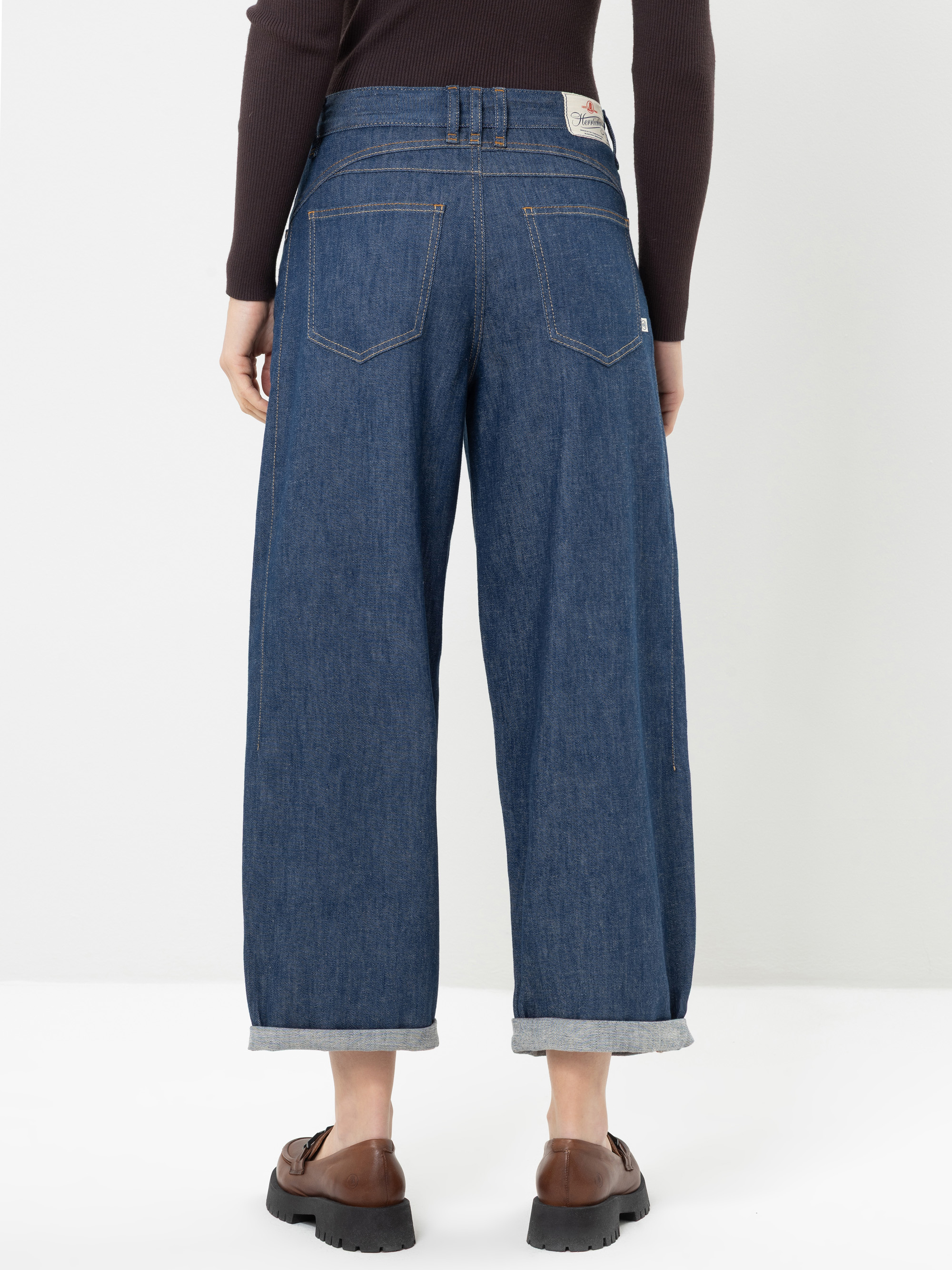 Eine Person steht mit dem Rücken zur Kamera und trägt eine blaue Jeans mit weitem Bein und umgeschlagenen Säumen, ein dunkles Langarm-Oberteil und braune Schuhe vor einem schlichten weißen Hintergrund.