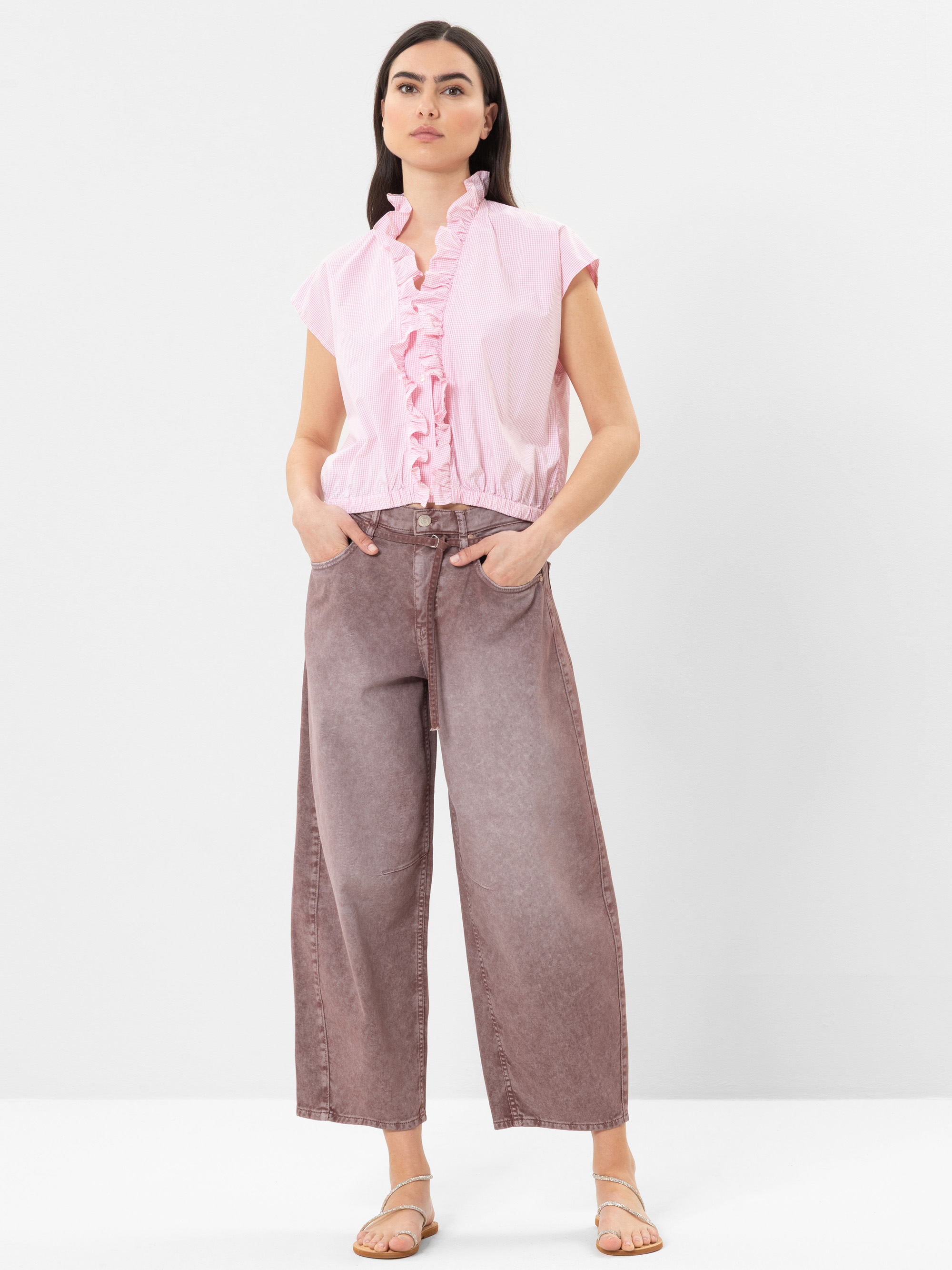 Eine Frau steht vor einem schlichten weißen Hintergrund und trägt eine hellrosa, kurzärmelige Rüschenbluse, eine weitbeinige lila Jeans und beigefarbene flache Sandalen, die Hände in den Taschen.
