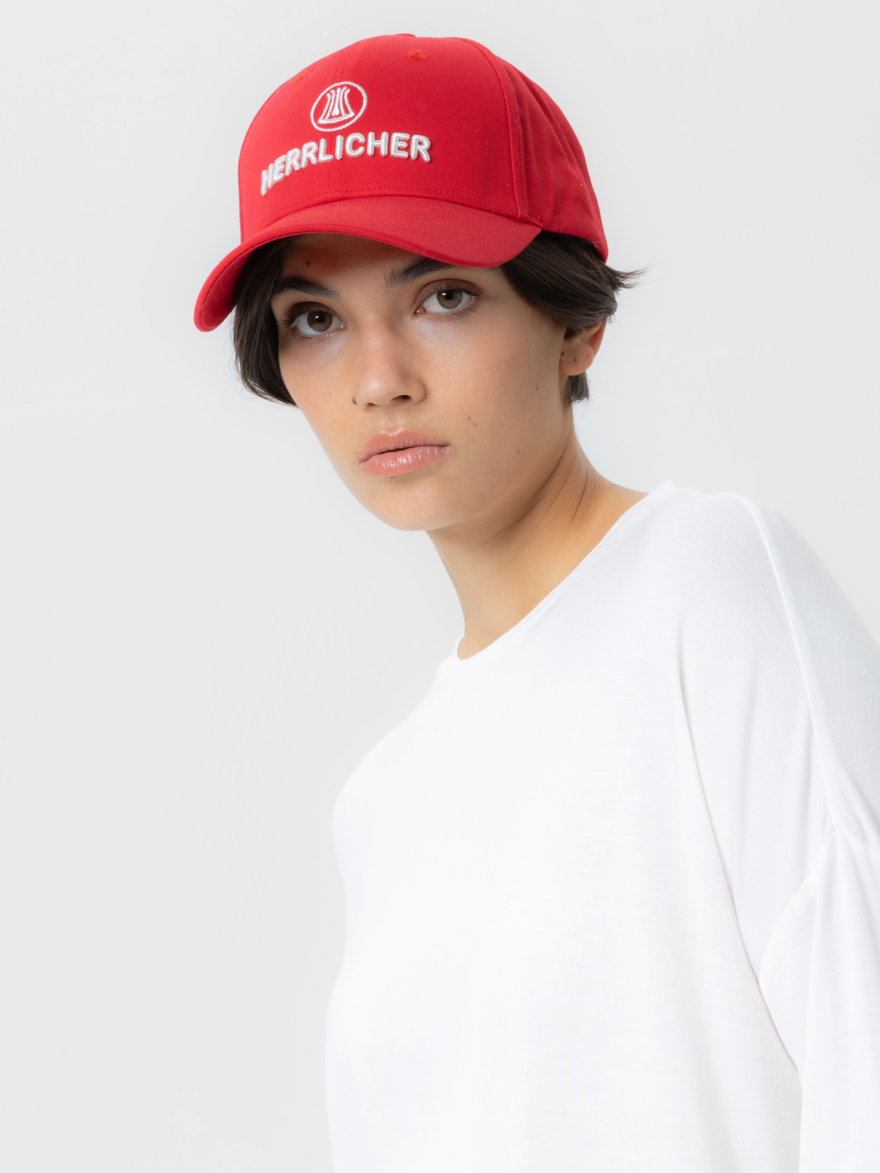 Eine Person mit kurzen dunklen Haaren, die eine rote Mütze mit der Aufschrift HERLLICHER und ein weißes Hemd trägt, steht vor einem schlichten hellen Hintergrund.