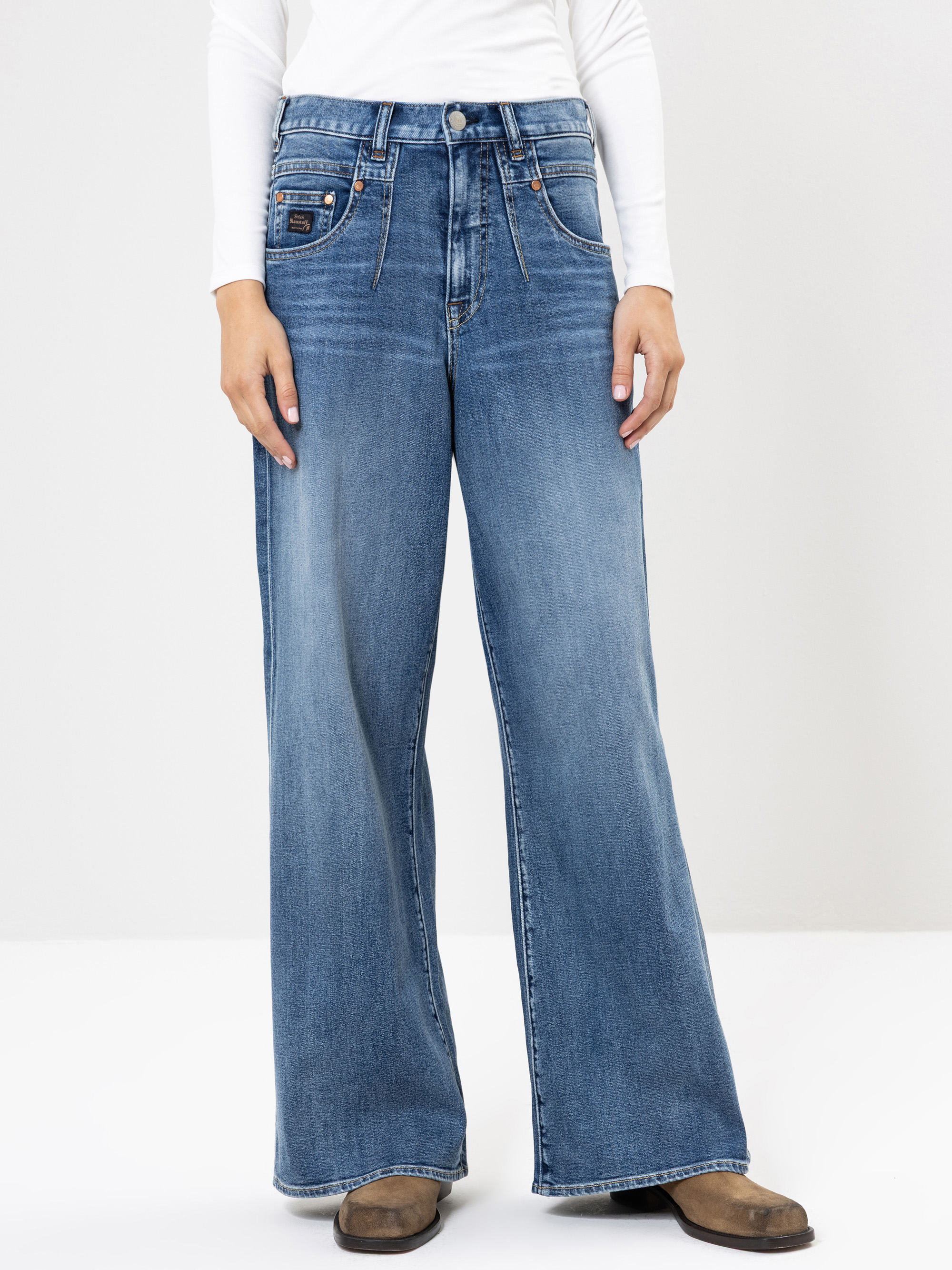 Eine Person, die eine blaue Jeans mit hoher Taille und weitem Bein, ein langärmeliges weißes Oberteil und braune Schuhe trägt, steht vor einem schlichten hellen Hintergrund. Der Kopf der Person ist auf dem Bild nicht zu sehen.