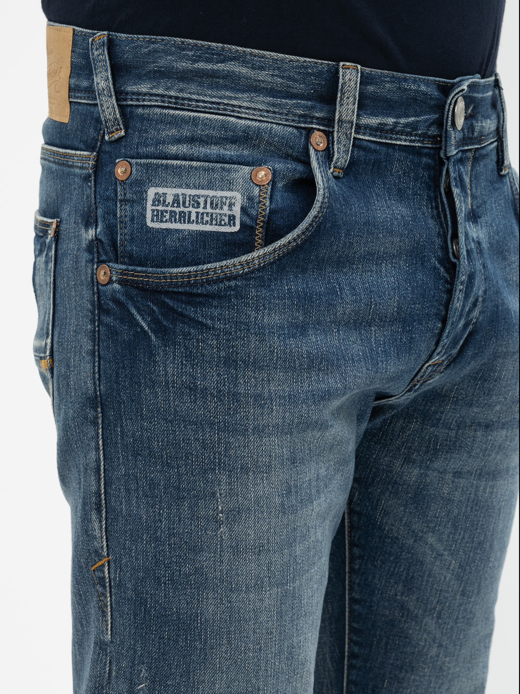 Nahaufnahme einer Person, die eine blaue Denim-Jeans und ein schwarzes Oberteil trägt. Die Jeans hat sichtbare Nähte, kupferne Nieten und ein kleines Etikett auf der Münztasche mit der Aufschrift BLAUSTOFF HERZLICHER.
