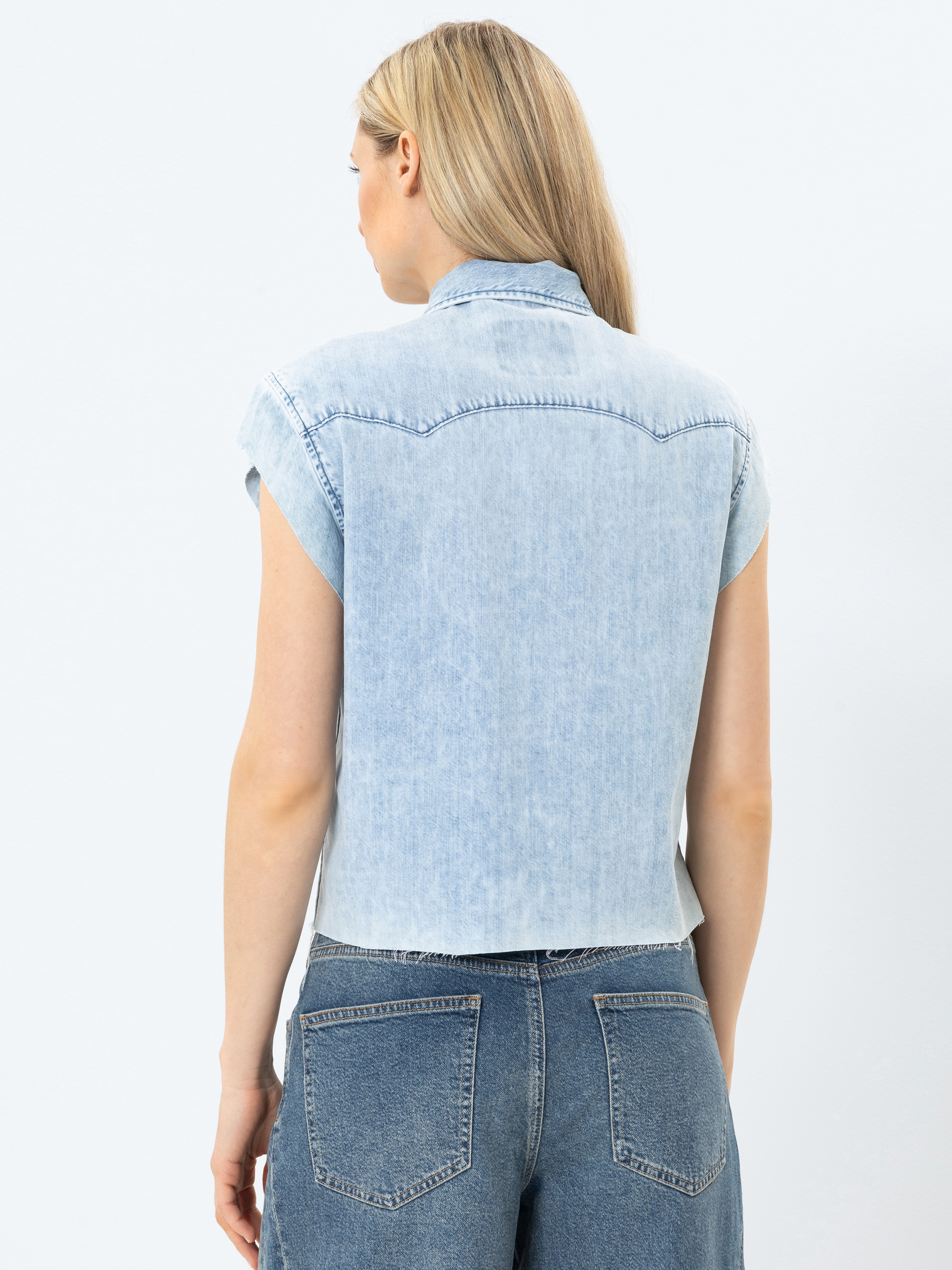 Eine Frau mit langen blonden Haaren trägt ein hellblaues, kurzärmeliges Jeanshemd und hochgeschnittene blaue Jeans, die von hinten vor einem schlichten weißen Hintergrund gezeigt werden.