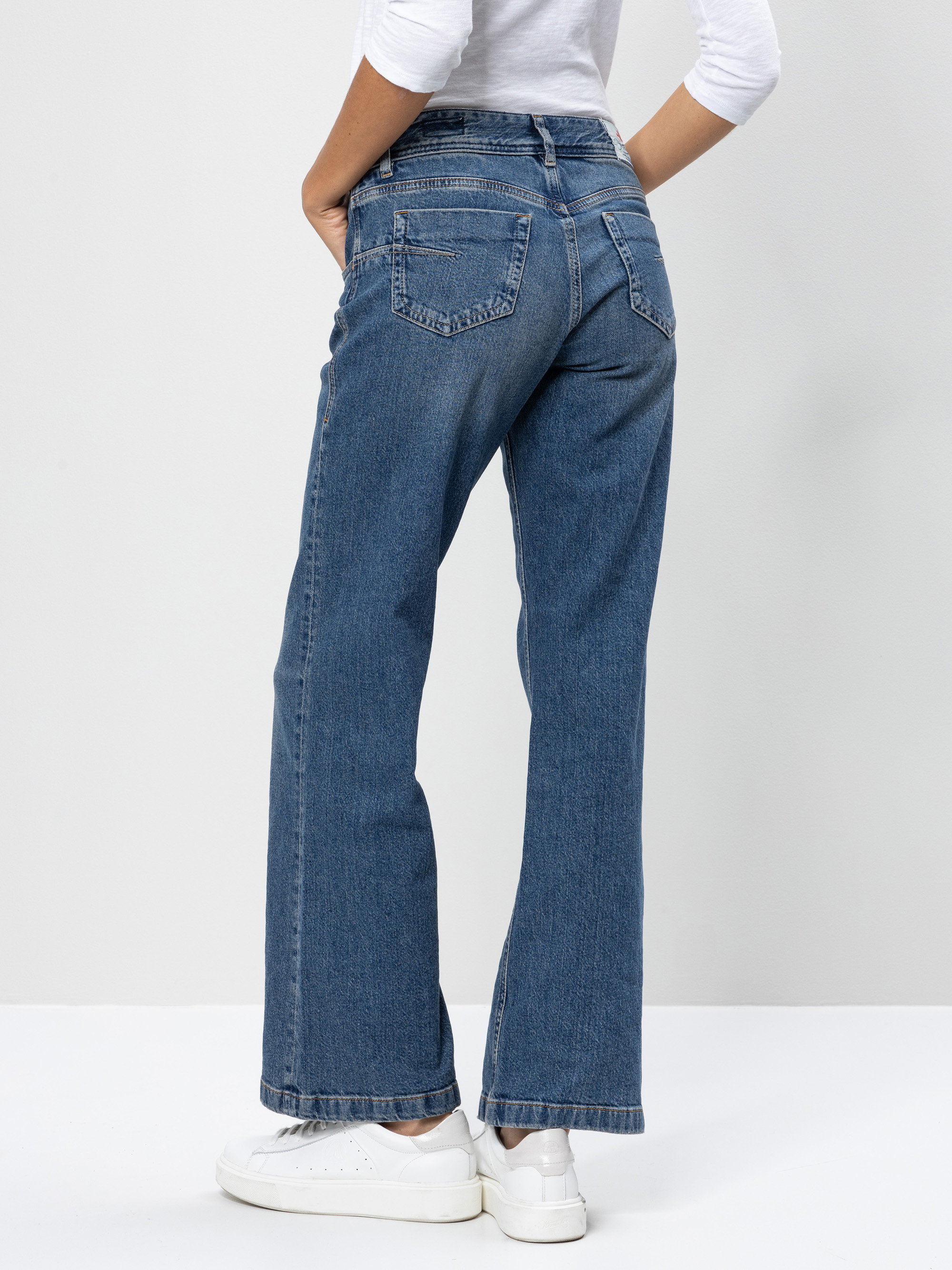 Person, die eine blaue Jeans mit geradem Bein, ein weißes langärmeliges Hemd und weiße Turnschuhe trägt, steht mit den Händen in den Hosentaschen und blickt von der Kamera weg in Richtung einer schlichten hellen Wand.