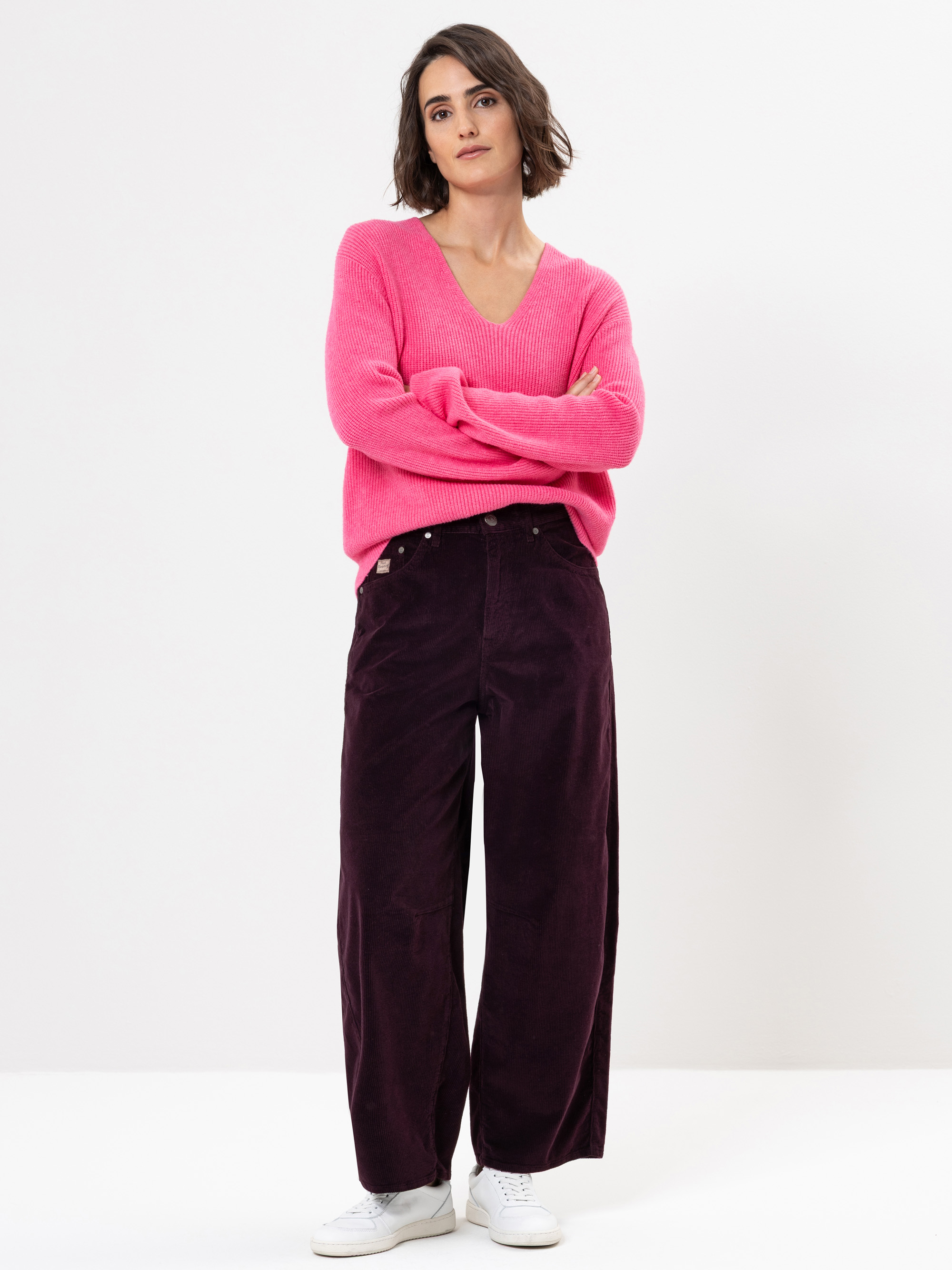 Eine Frau mit kurzen braunen Haaren steht vor einem weißen Hintergrund. Sie trägt einen hellrosa Pullover, eine weite dunkelviolette Hose und weiße Turnschuhe, hat die Arme verschränkt und macht einen entspannten Eindruck.