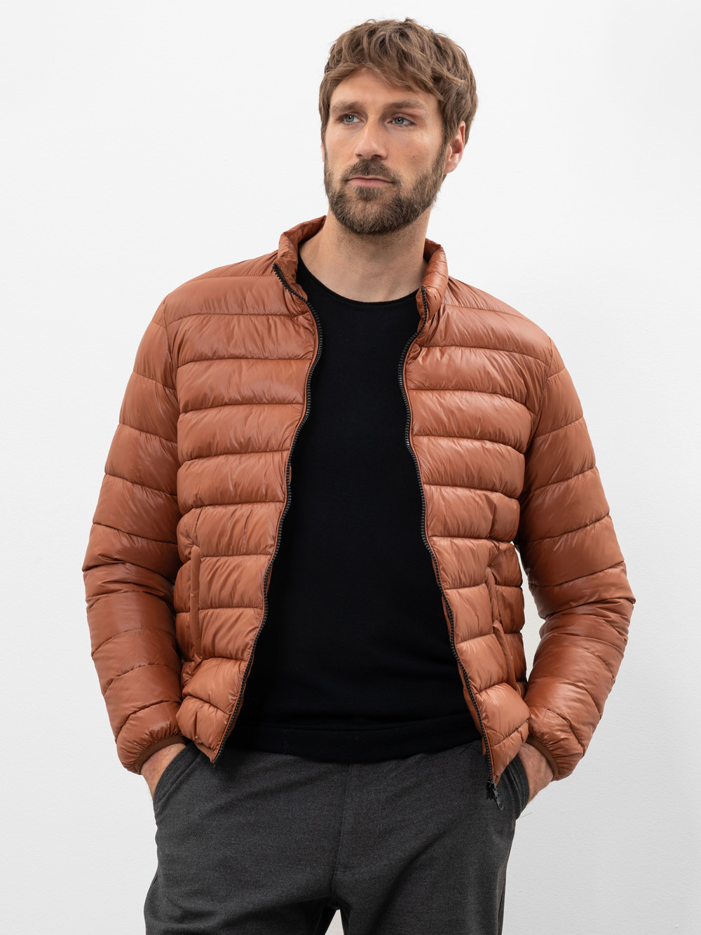 Ein Mann mit kurzen braunen Haaren und einem Bart trägt eine rostfarbene Pufferjacke über einem schwarzen Hemd und einer grauen Hose und steht mit den Händen in den Taschen vor einem weißen Hintergrund.