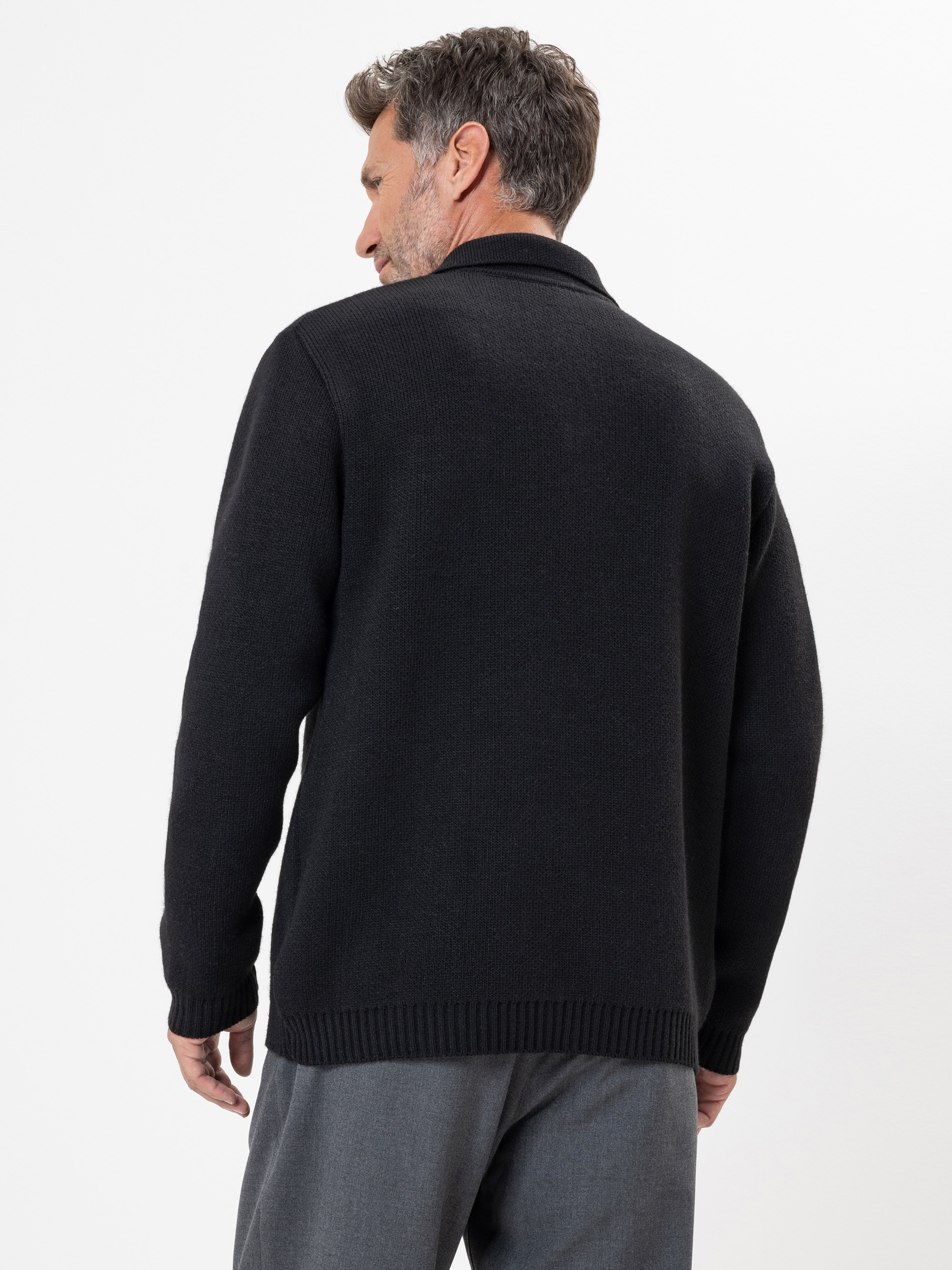 Ein Mann mit kurzen braunen Haaren steht mit dem Rücken zur Kamera, trägt einen schwarzen Pullover und eine graue Hose vor einem schlichten weißen Hintergrund.