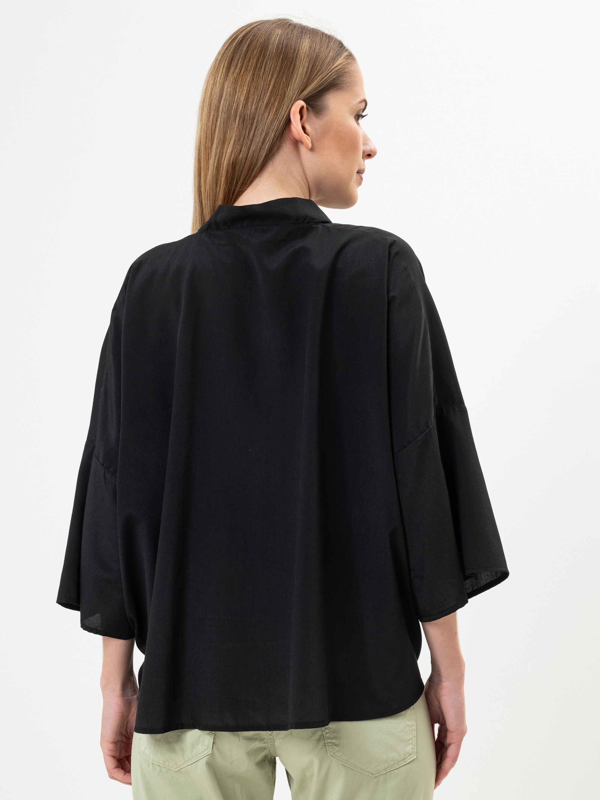 Eine Frau mit glattem, hellbraunem Haar trägt ein lockeres schwarzes, langärmeliges Oberteil und eine hellgrüne Hose und steht mit dem Rücken zur Kamera vor einem schlichten weißen Hintergrund.