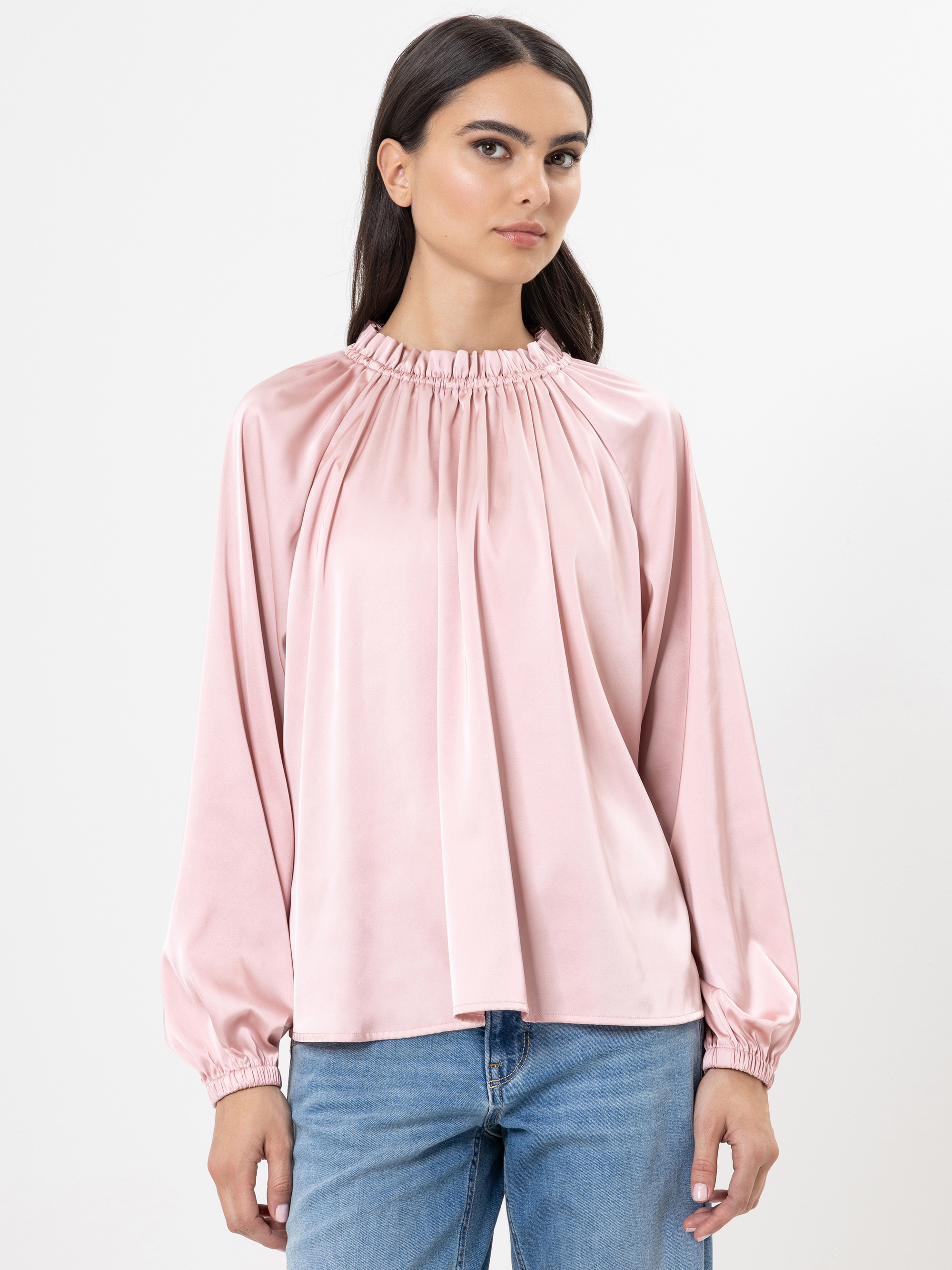 Eine Frau mit langen dunklen Haaren trägt eine lockere, hellrosa Satinbluse mit gerafftem Ausschnitt und langen Ärmeln, gepaart mit blauen Jeans, vor einem schlichten weißen Hintergrund.