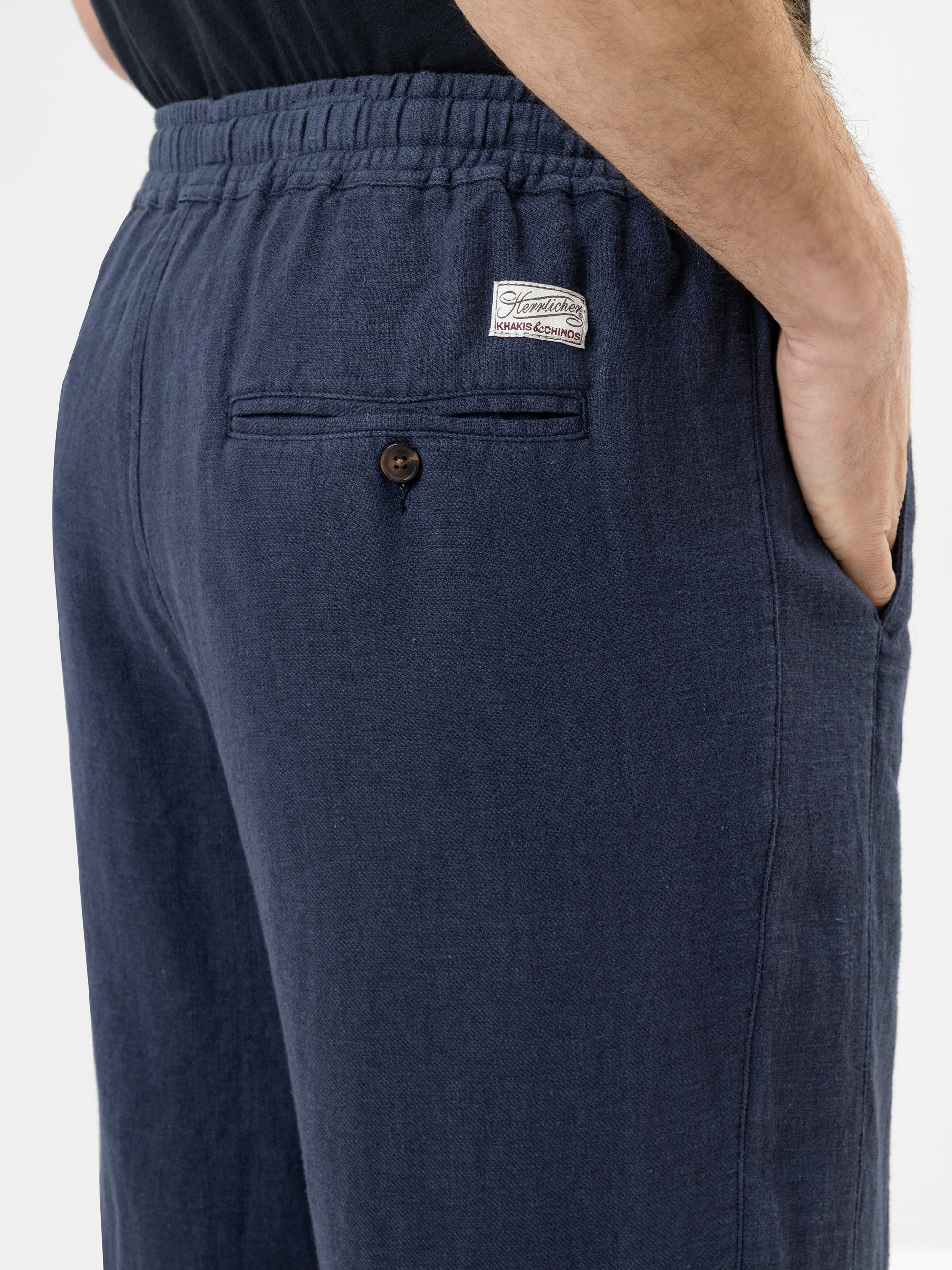 Eine Person, die eine marineblaue Hose mit einem elastischen Bund und einer Hand in der Tasche trägt; darüber befindet sich eine Gesäßtasche mit einem Knopf und einem kleinen weißen Etikett.