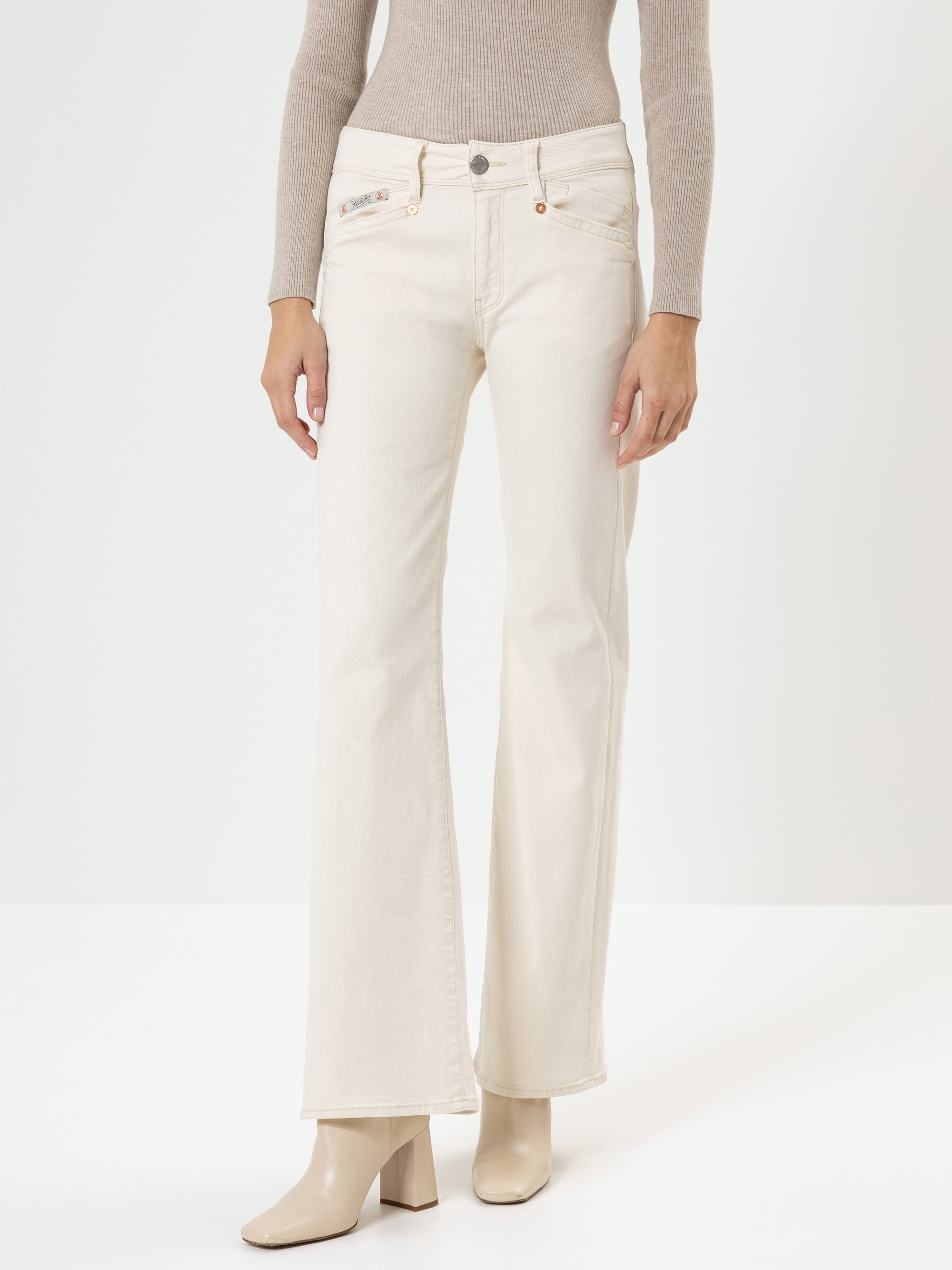 Eine Person, die cremefarbene Bootcut-Jeans, ein beigefarbenes langärmeliges Rippenoberteil und cremefarbene Stiefeletten trägt, steht vor einem schlichten weißen Hintergrund. Nur der Körper von den Schultern abwärts ist sichtbar.