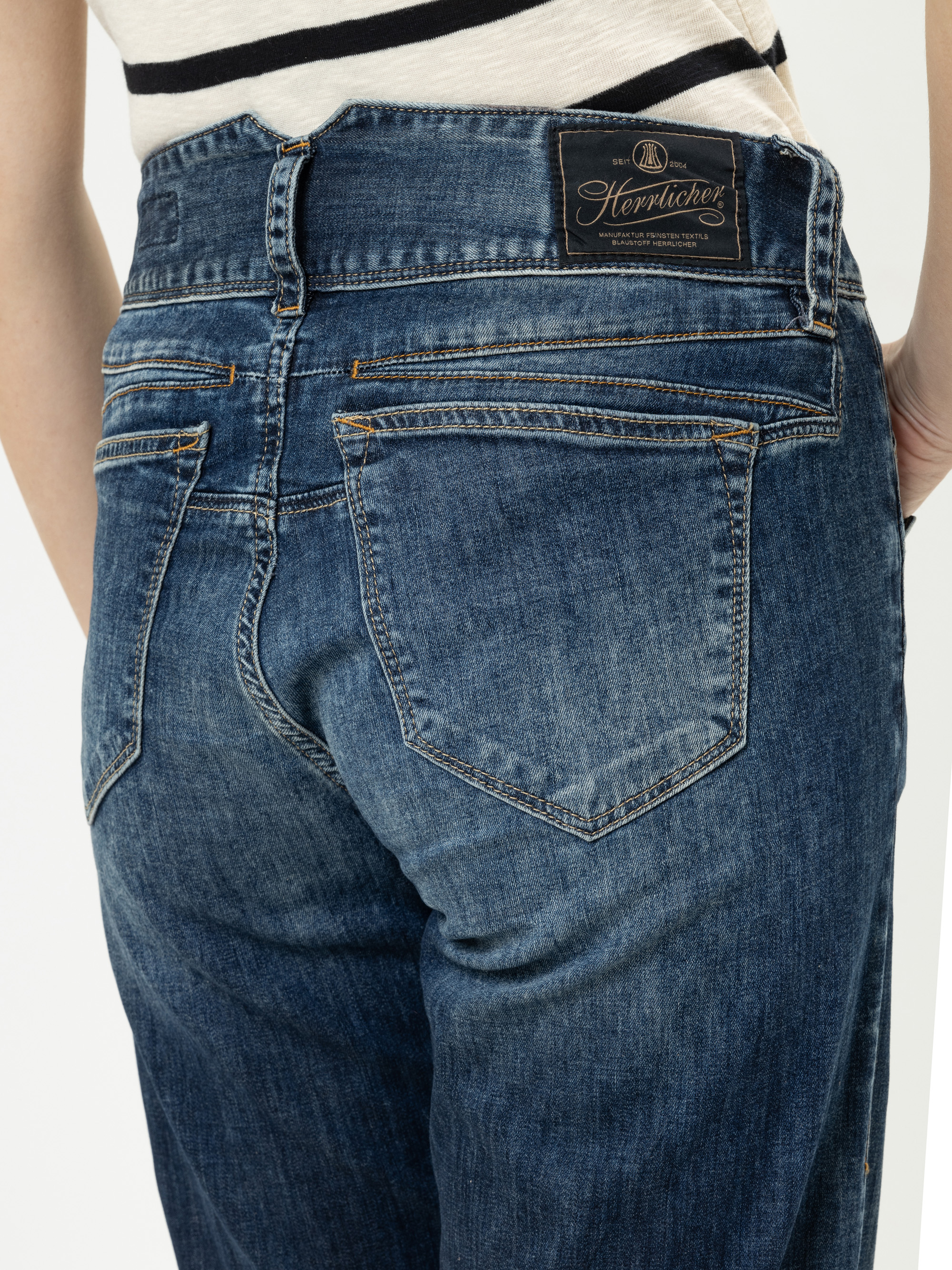 Nahaufnahme einer Person, die eine blaue Denim-Jeans mit sichtbaren Gesäßtaschen und einem schwarzen Herrlicher-Markenetikett am Bund trägt. Die Person steht vor einem schlichten, hellen Hintergrund.