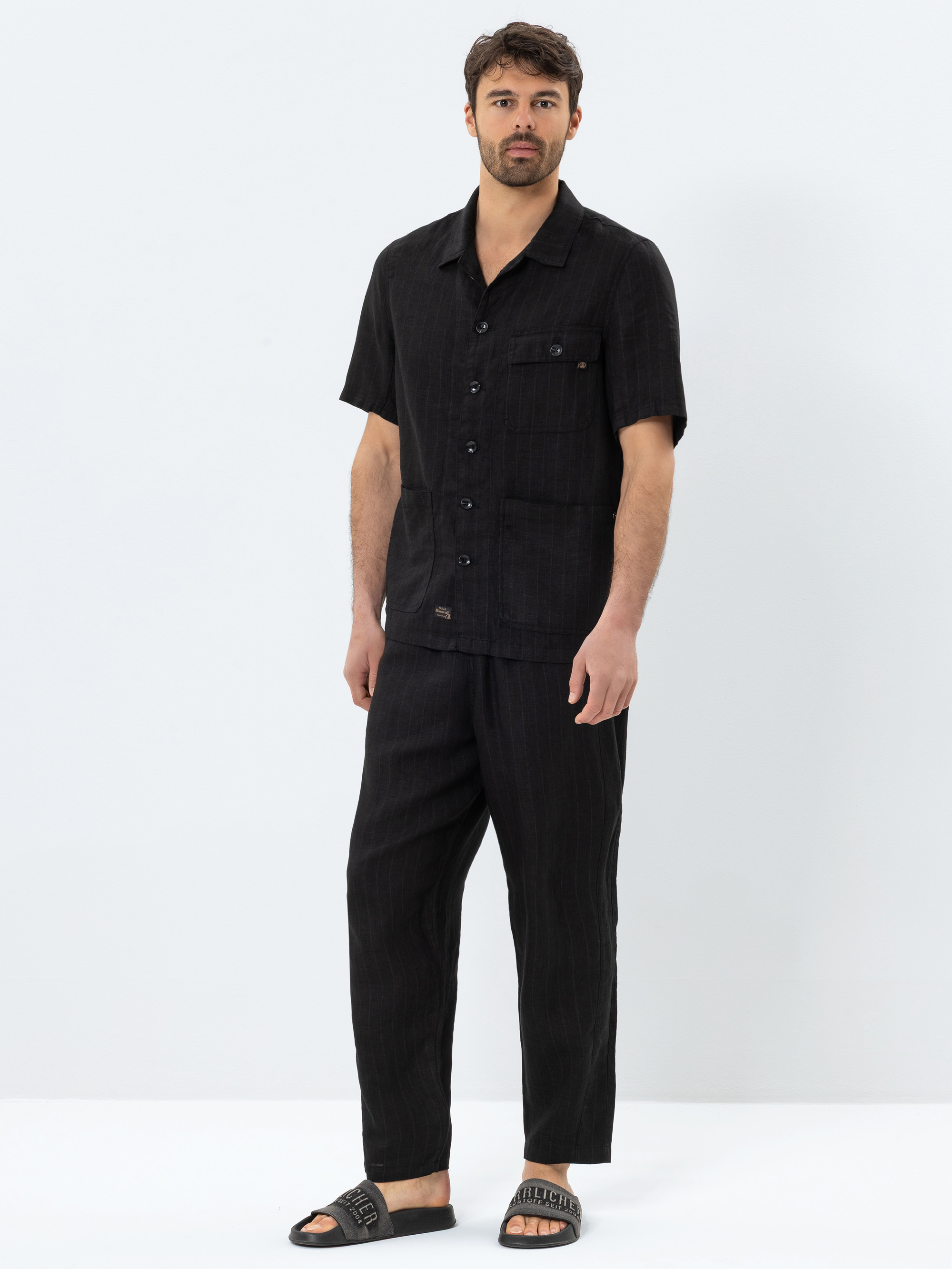 Ein Mann mit kurzen dunklen Haaren und einem Bart trägt ein schwarzes, kurzärmeliges Hemd zum Aufknöpfen, eine dazu passende schwarze Hose und schwarze Slide-Sandalen mit einem Schriftzug auf den Riemen, die vor einem schlichten weißen Hintergrund ste