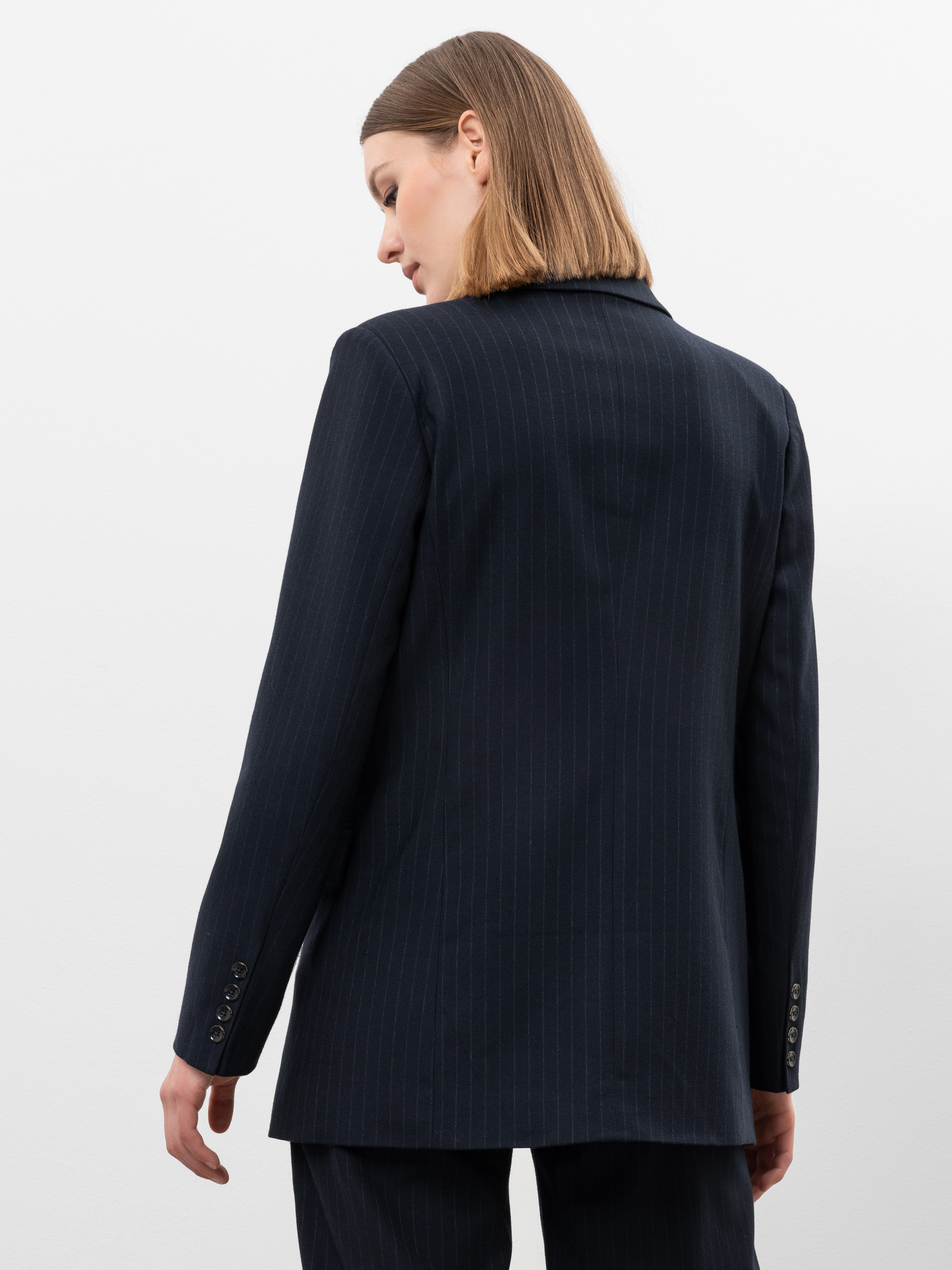 Eine Person mit glattem, schulterlangem Haar trägt einen dunklen Nadelstreifenanzug und steht mit dem Blick über die linke Schulter vor einem schlichten weißen Hintergrund.