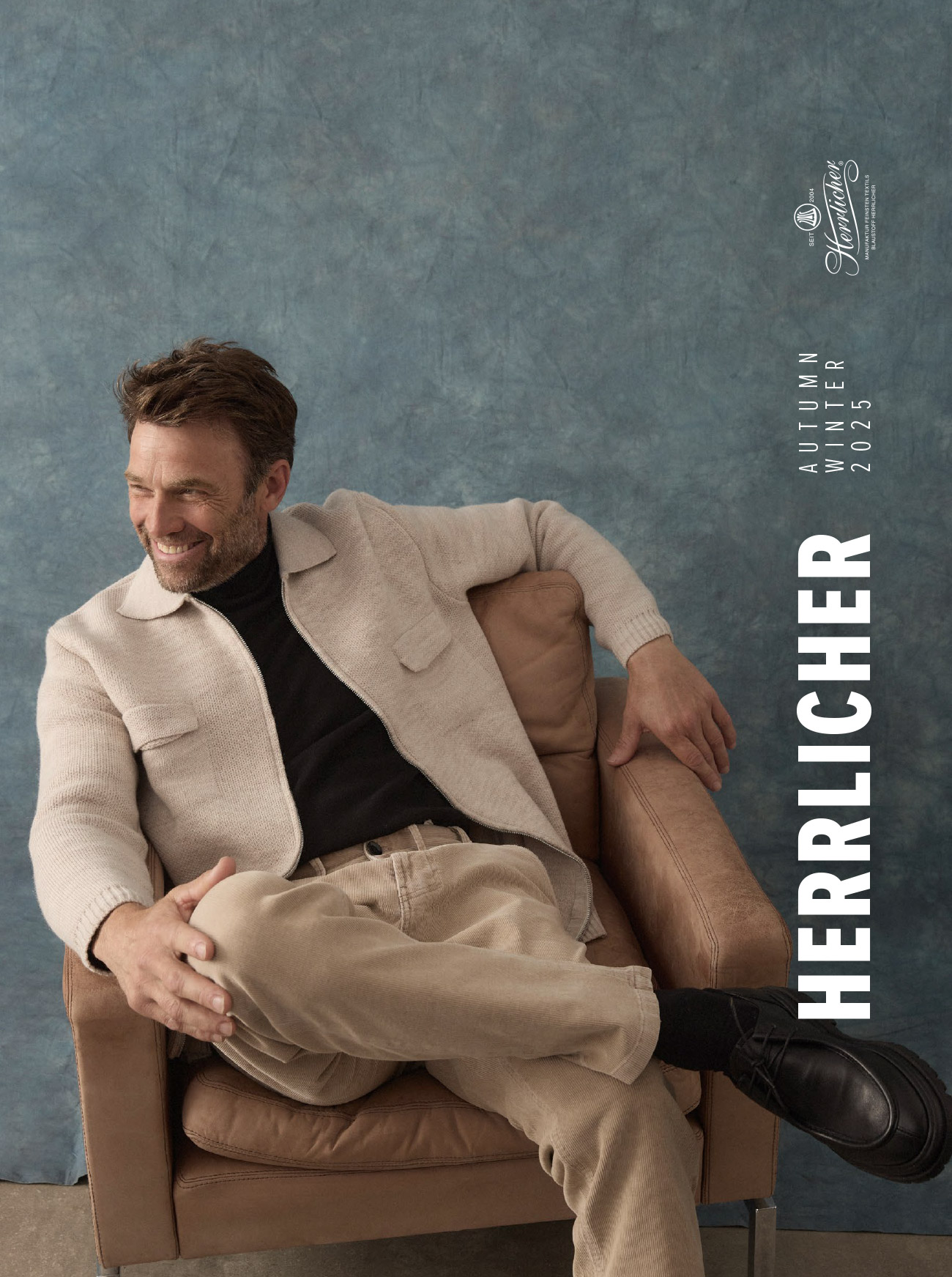 Ein Mann sitzt in einem hellbraunen Sessel, lächelt und blickt zur Seite. Er trägt eine helle Jacke, einen schwarzen Rollkragenpullover, eine beige Hose und schwarze Schuhe. Der Hintergrund ist blau mit dem Text HERRLICHER AUTUMN WINTER 2025 auf der rec