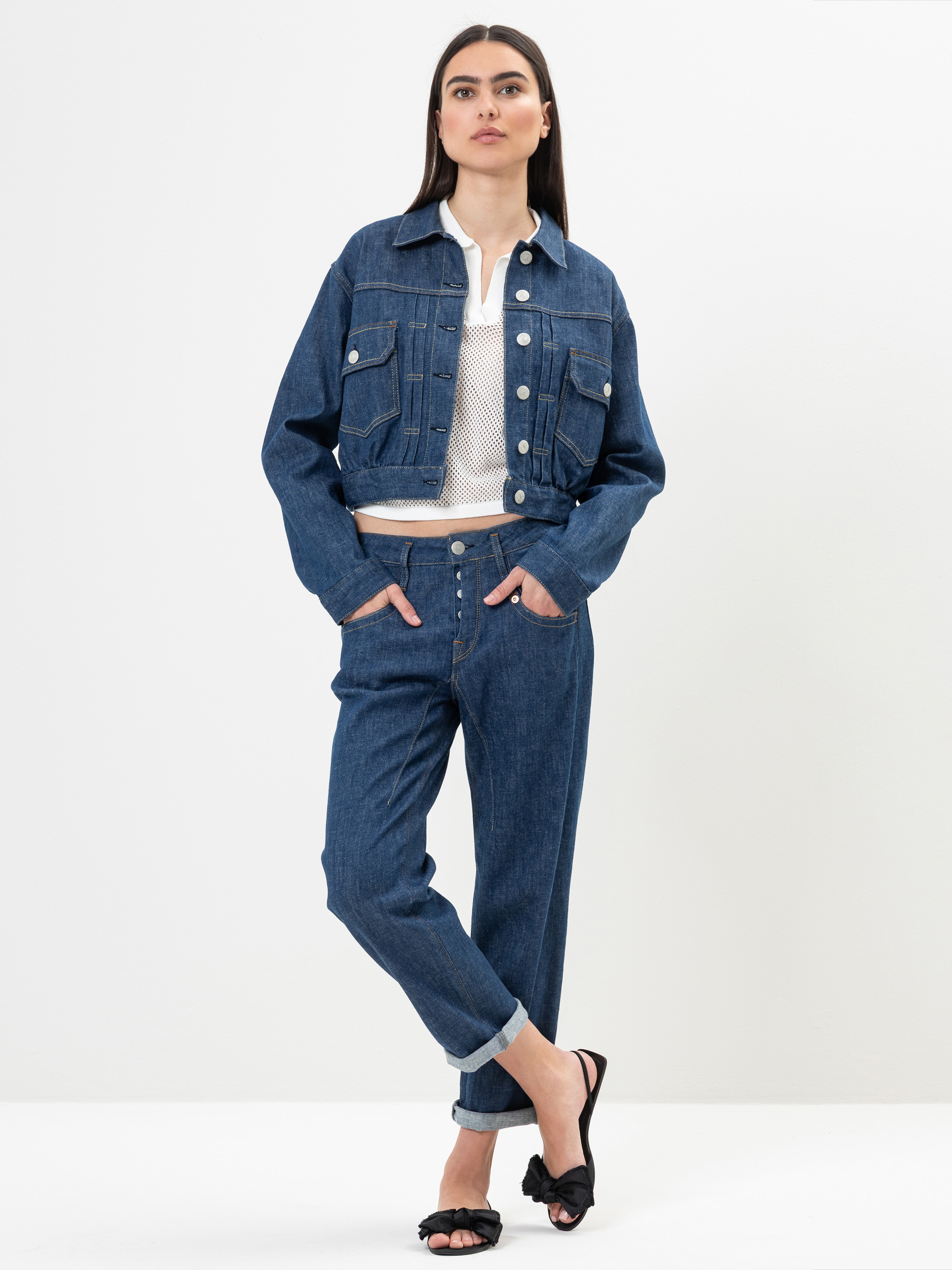 Eine Frau steht vor einem schlichten weißen Hintergrund und trägt eine abgeschnittene Jeansjacke, ein weißes Oberteil, hochgeschnittene Jeans mit Bündchen und schwarze Sandalen. Sie hat glattes dunkles Haar und ihre Hände stecken in den Taschen.