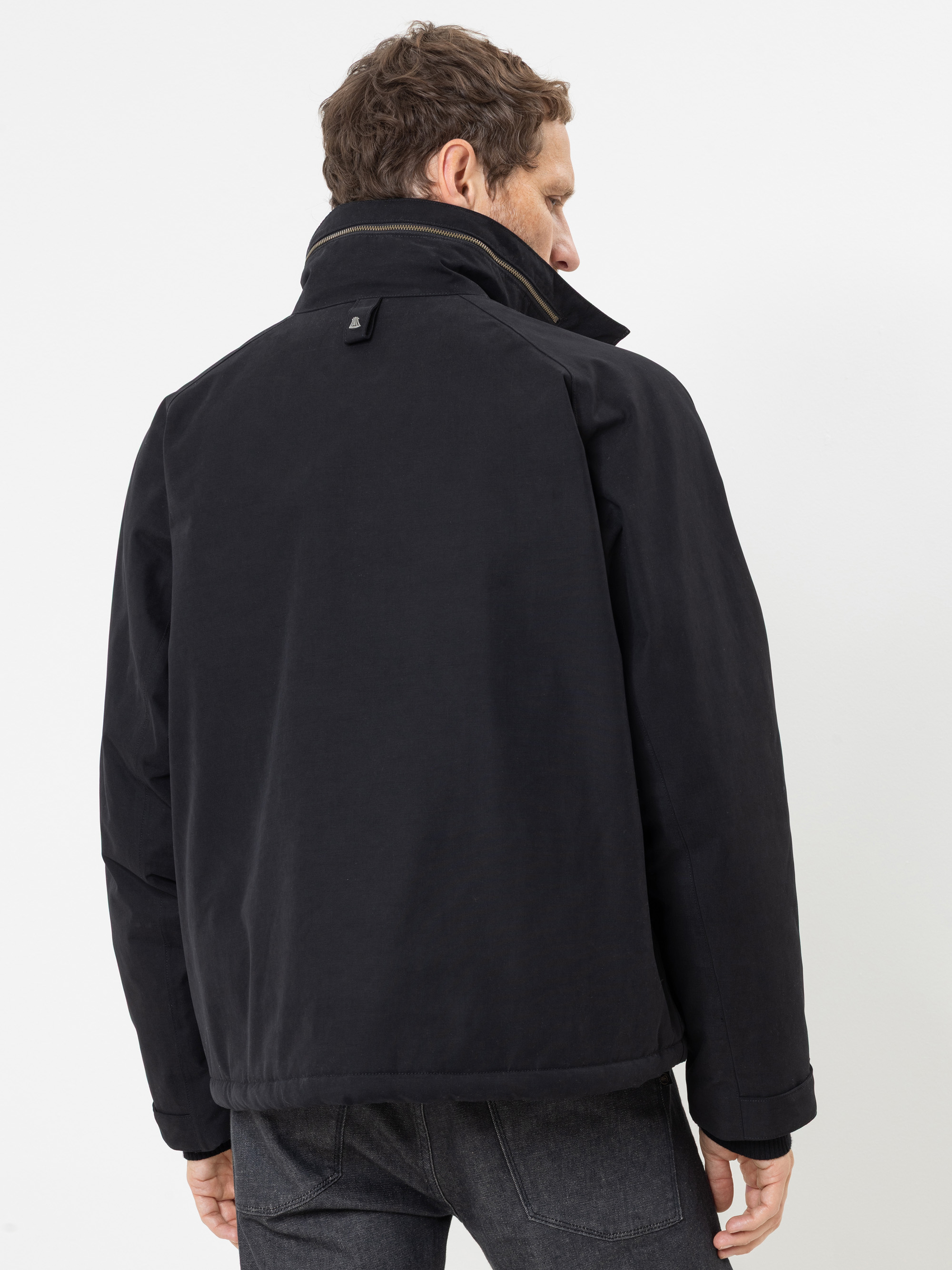 Ein Mann mit kurzen braunen Haaren, der eine schwarze Jacke und dunkle Jeans trägt, steht mit dem Rücken zur Kamera und blickt nach rechts vor einem schlichten weißen Hintergrund.