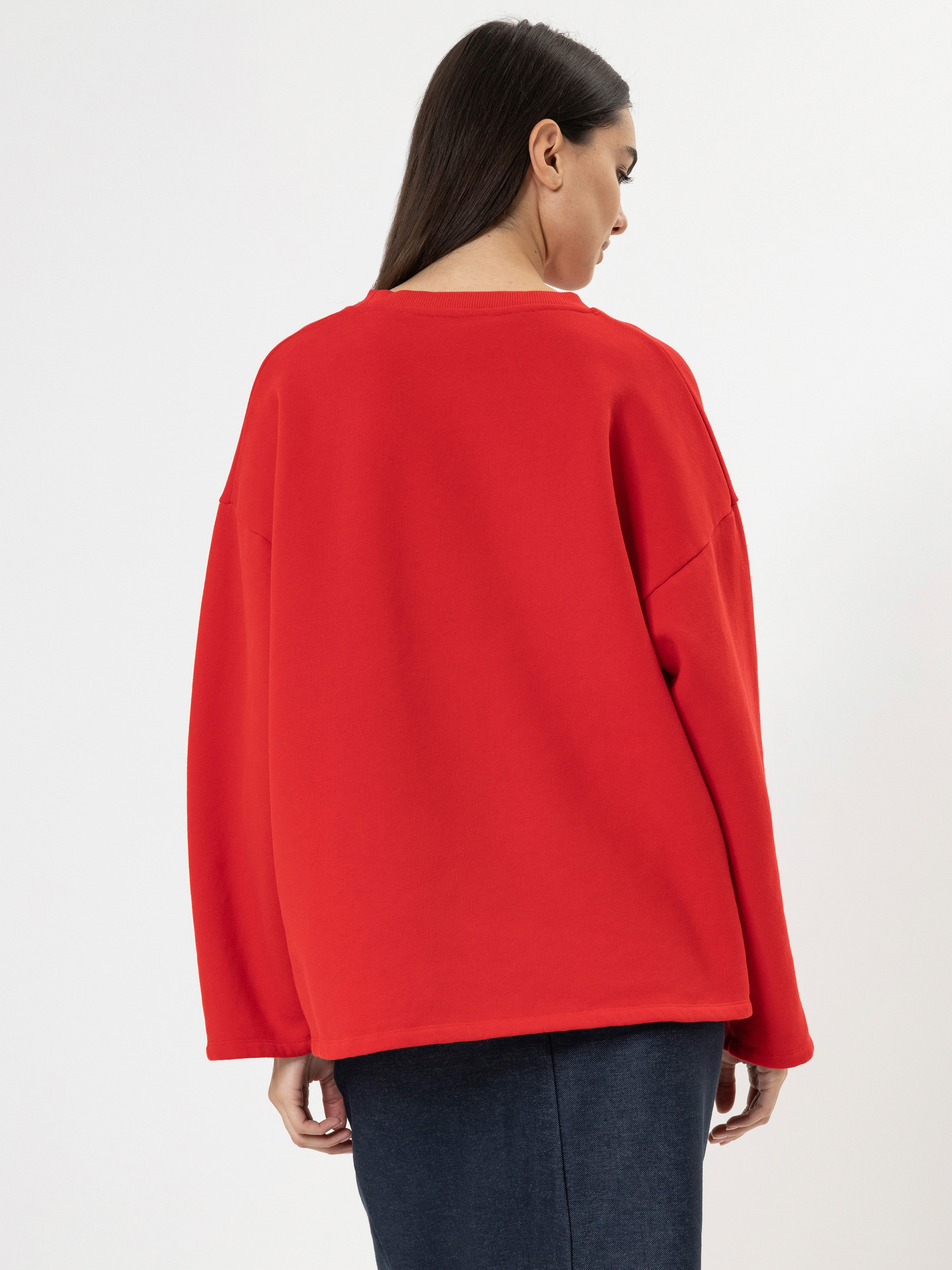 Eine Frau mit langen dunklen Haaren wird von hinten gezeigt. Sie trägt einen weiten roten Pullover und einen dunklen Rock und steht vor einem schlichten weißen Hintergrund.