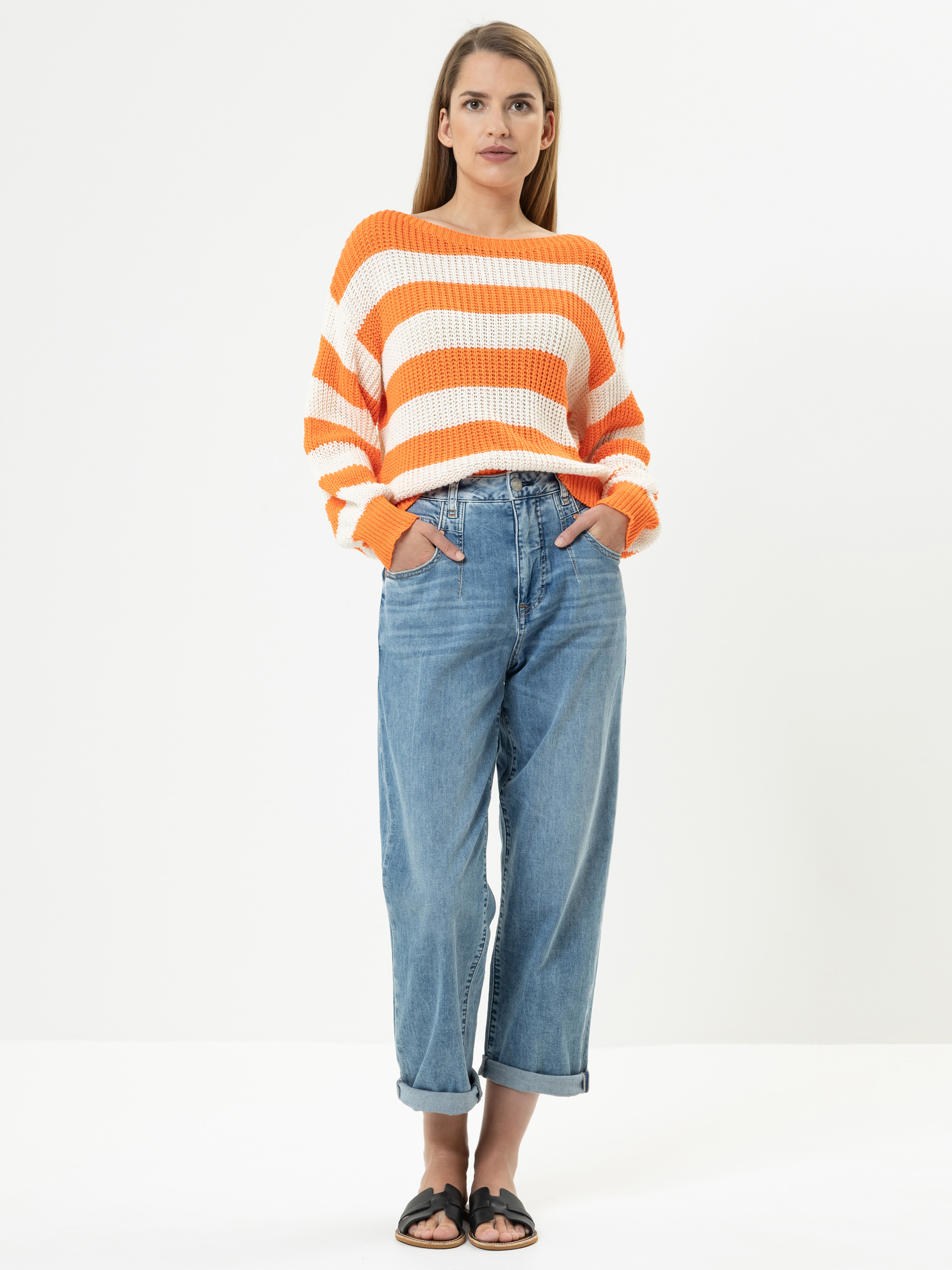 Eine Frau steht mit dem Gesicht nach vorne, trägt einen lockeren orange-weiß gestreiften Pullover, eine blaue Jeans mit hohem Bund und schwarzen Sandalen, die Hände in den Taschen, vor einem schlichten weißen Hintergrund.