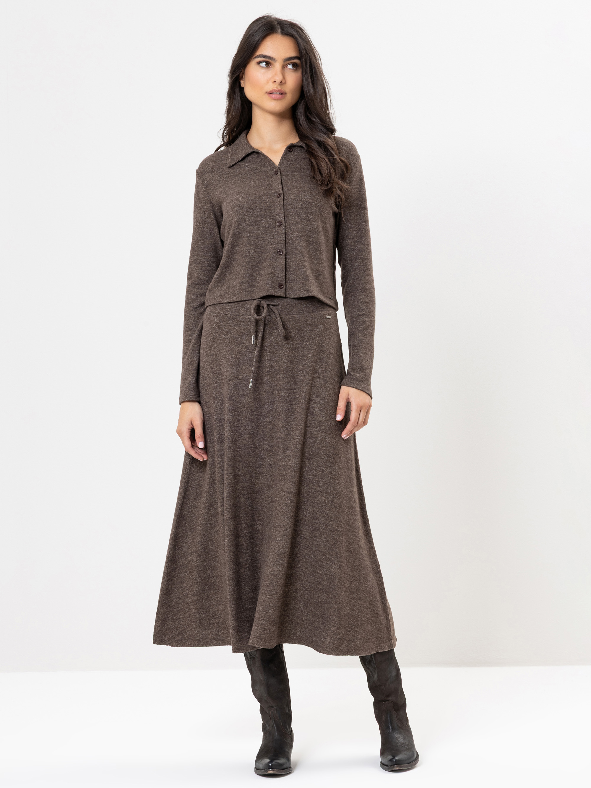 Eine Frau steht vor einem schlichten weißen Hintergrund und trägt ein braunes, langärmeliges Oberteil mit Knöpfen und einen dazu passenden knöchellangen Rock mit Kordelzug in der Taille, gepaart mit dunklen, kniehohen Stiefeln.