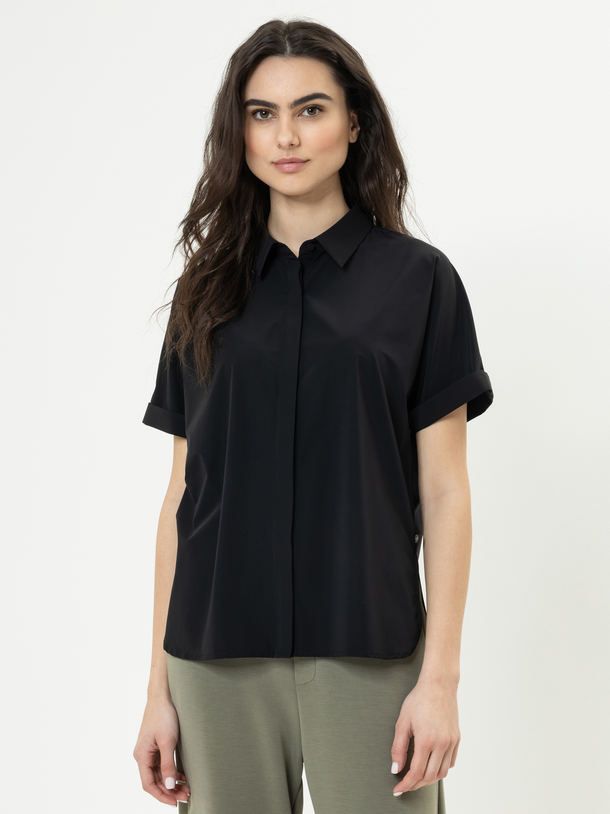 Eine Frau mit langen, gewellten braunen Haaren trägt ein locker sitzendes schwarzes, kurzärmeliges Button-up-Hemd und eine olivgrüne Hose und steht vor einem schlichten weißen Hintergrund.
