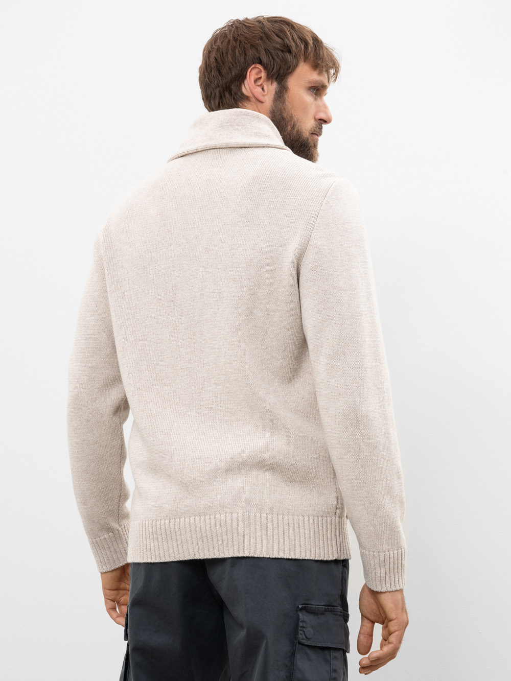 Ein Mann mit Bart, von hinten gesehen, trägt einen hellbeigen Strickpullover mit hohem Kragen und eine schwarze Cargohose und steht vor einem schlichten weißen Hintergrund.