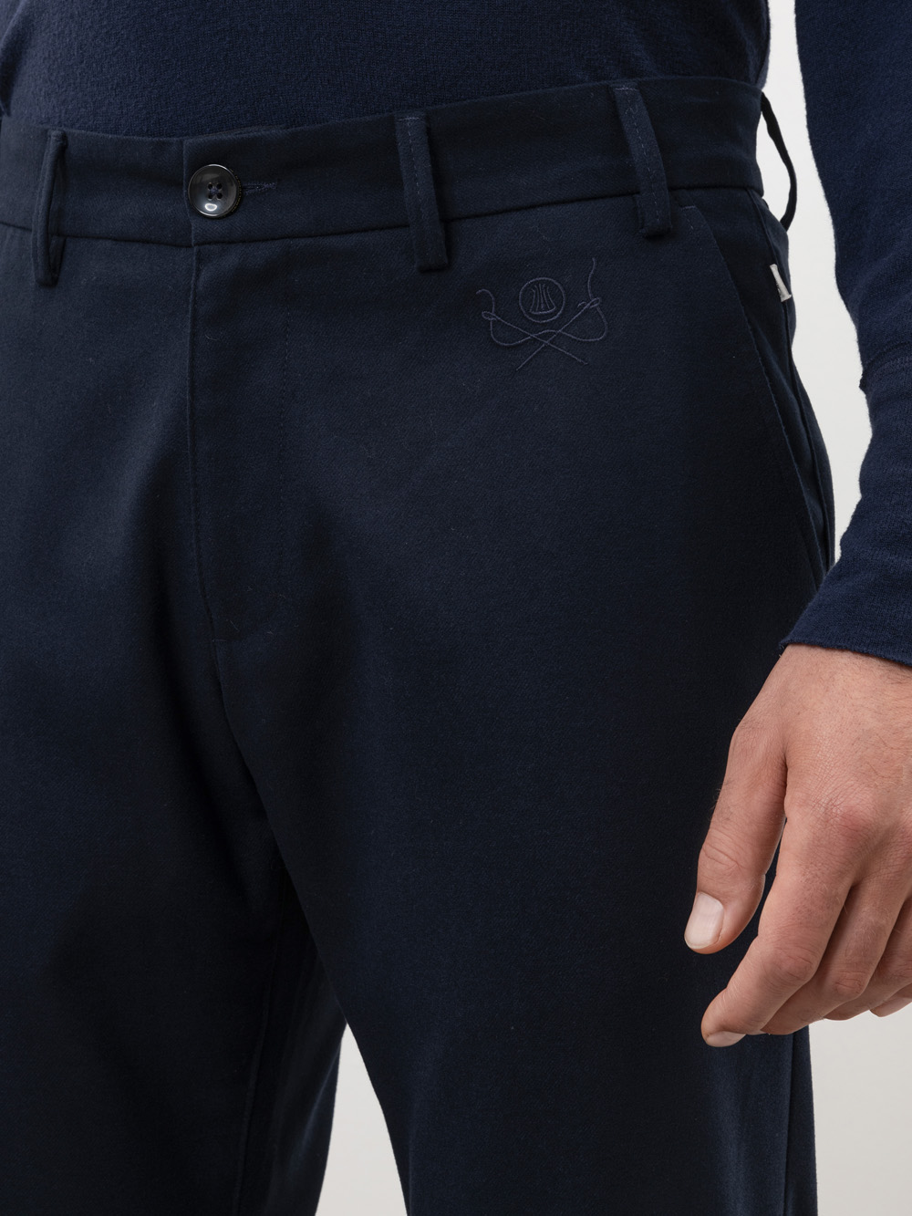 Eine Person, die eine dunkle, marineblaue Hose und ein passendes langärmeliges Hemd trägt. Die Hose hat einen Knopfverschluss, Gürtelschlaufen und ein gesticktes Wappen in der Nähe der rechten Tasche. Nur der untere Teil des Oberkörpers und die recht