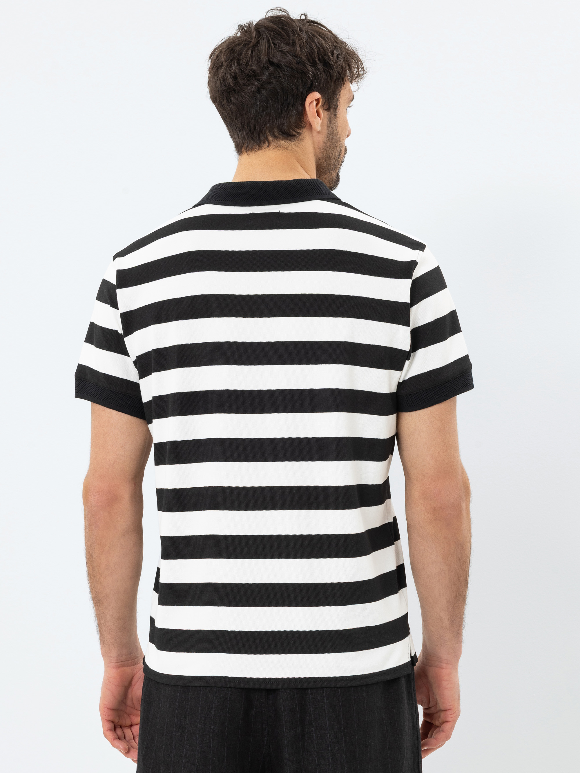 Ein Mann steht mit dem Rücken zur Kamera, trägt ein kurzärmeliges, schwarz-weißes, horizontal gestreiftes Polohemd und eine schwarze Hose vor einem schlichten weißen Hintergrund.