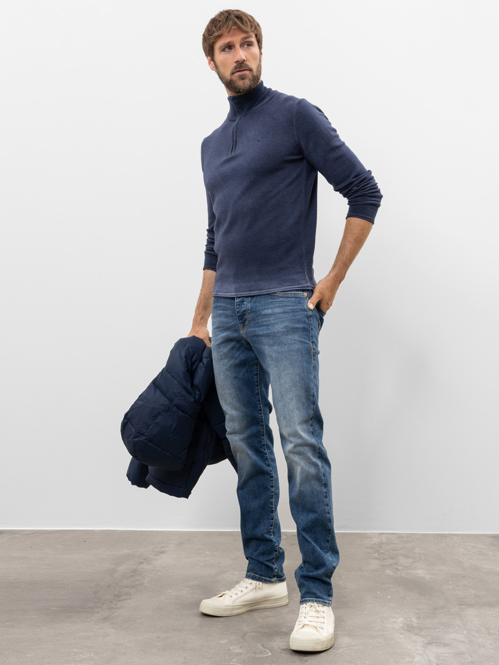 Ein Mann, der einen marineblauen Rollkragenpullover, blaue Jeans und weiße Turnschuhe trägt, steht auf einem grauen Boden, hält eine blaue Pufferjacke in einer Hand und blickt leicht zur Seite vor einem schlichten weißen Hintergrund.