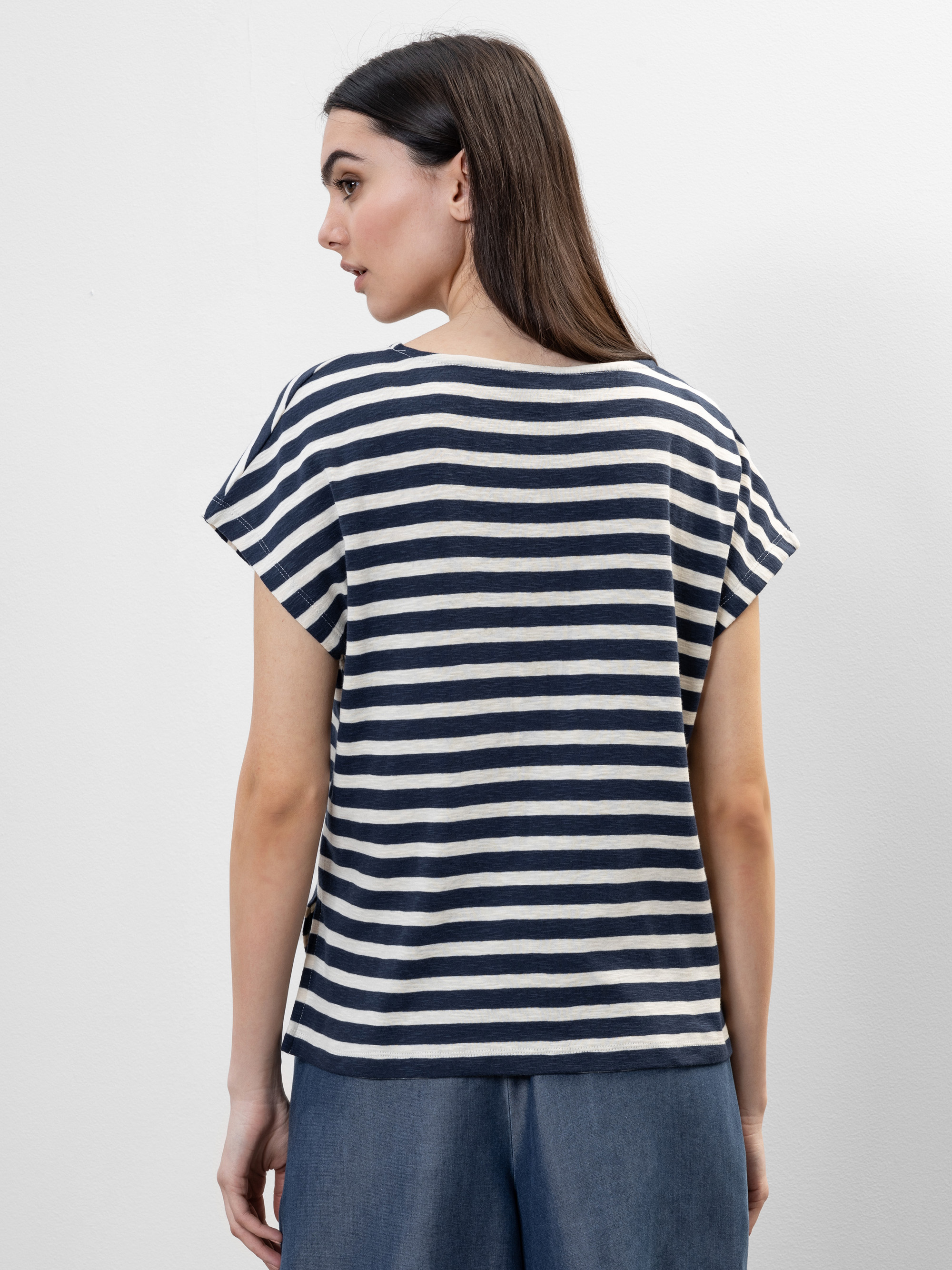 Frau mit langen braunen Haaren, die ein marineblau-weiß gestreiftes, kurzärmeliges Oberteil und eine blaue Hose trägt. Sie steht vor einem schlichten, hellen Hintergrund und schaut leicht von der Kamera weg.