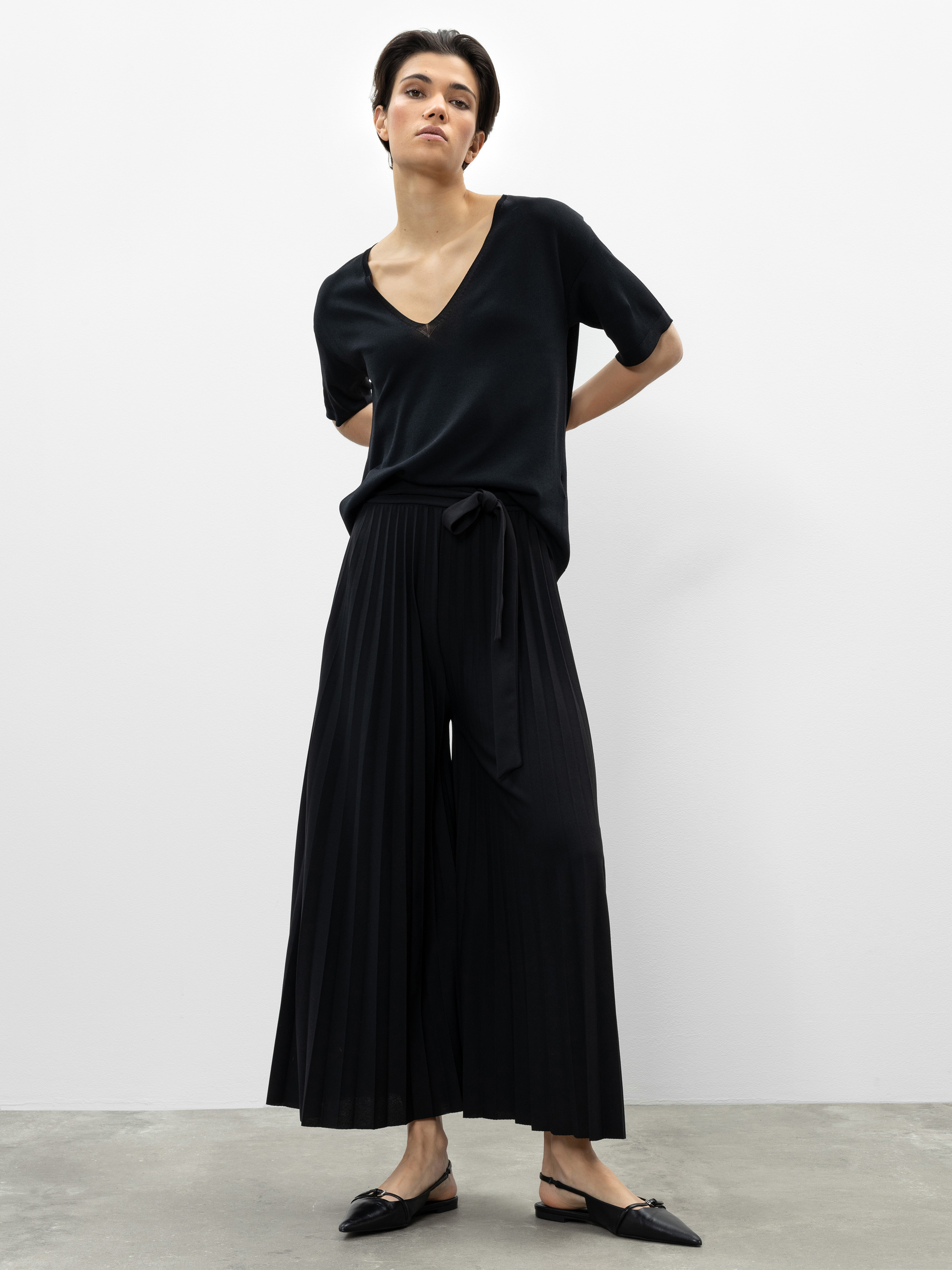 Eine Person steht vor einer schlichten weißen Wand. Sie trägt ein lockeres schwarzes Oberteil mit V-Ausschnitt, eine schwarze, in Falten gelegte Hose mit weitem Bein und einer Krawatte in der Taille sowie schwarze, spitze, flache Schuhe. Ihr Haar ist ku