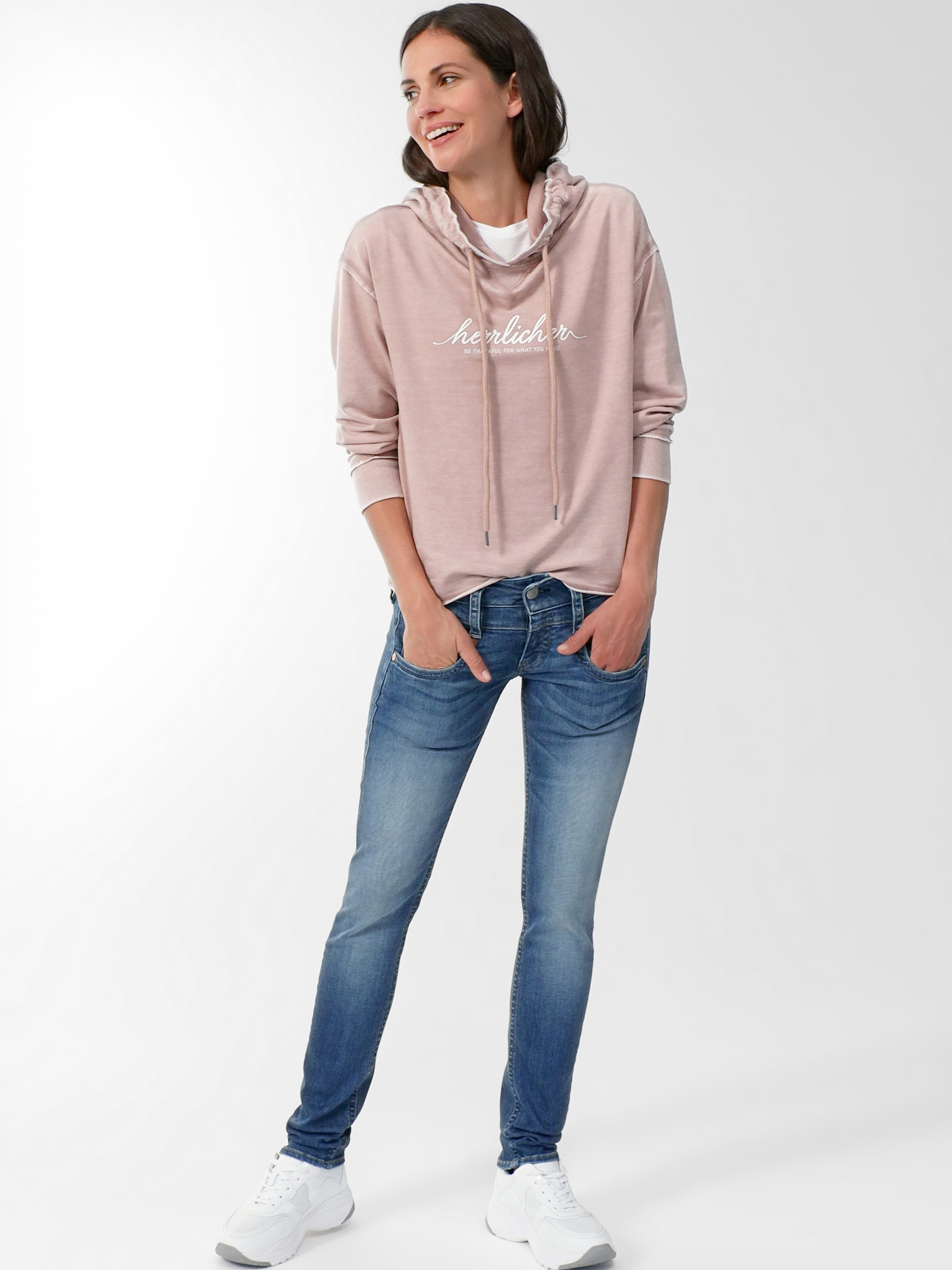 Eine Frau steht lächelnd mit den Händen in den Jeanstaschen da. Sie trägt einen hellrosa Kapuzenpullover mit weißer Aufschrift, weiße Turnschuhe und ein weißes T-Shirt vor einem schlichten hellen Hintergrund.