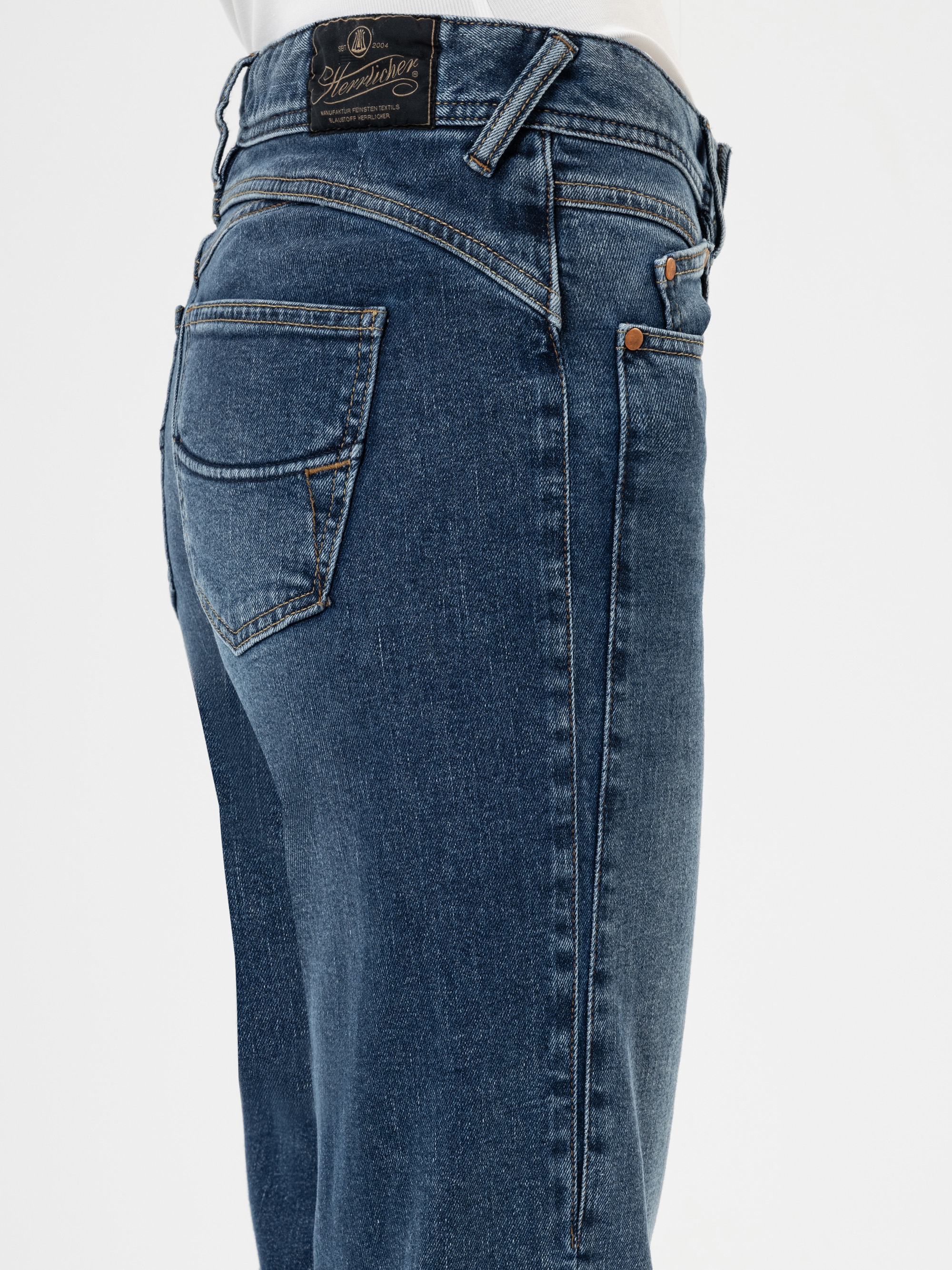 Nahaufnahme einer Person, die eine blaue Denim-Jeans trägt, mit Gesäßtasche, Gürtelschlaufen und einem schwarzen Markenetikett am Bund vor einem schlichten weißen Hintergrund.