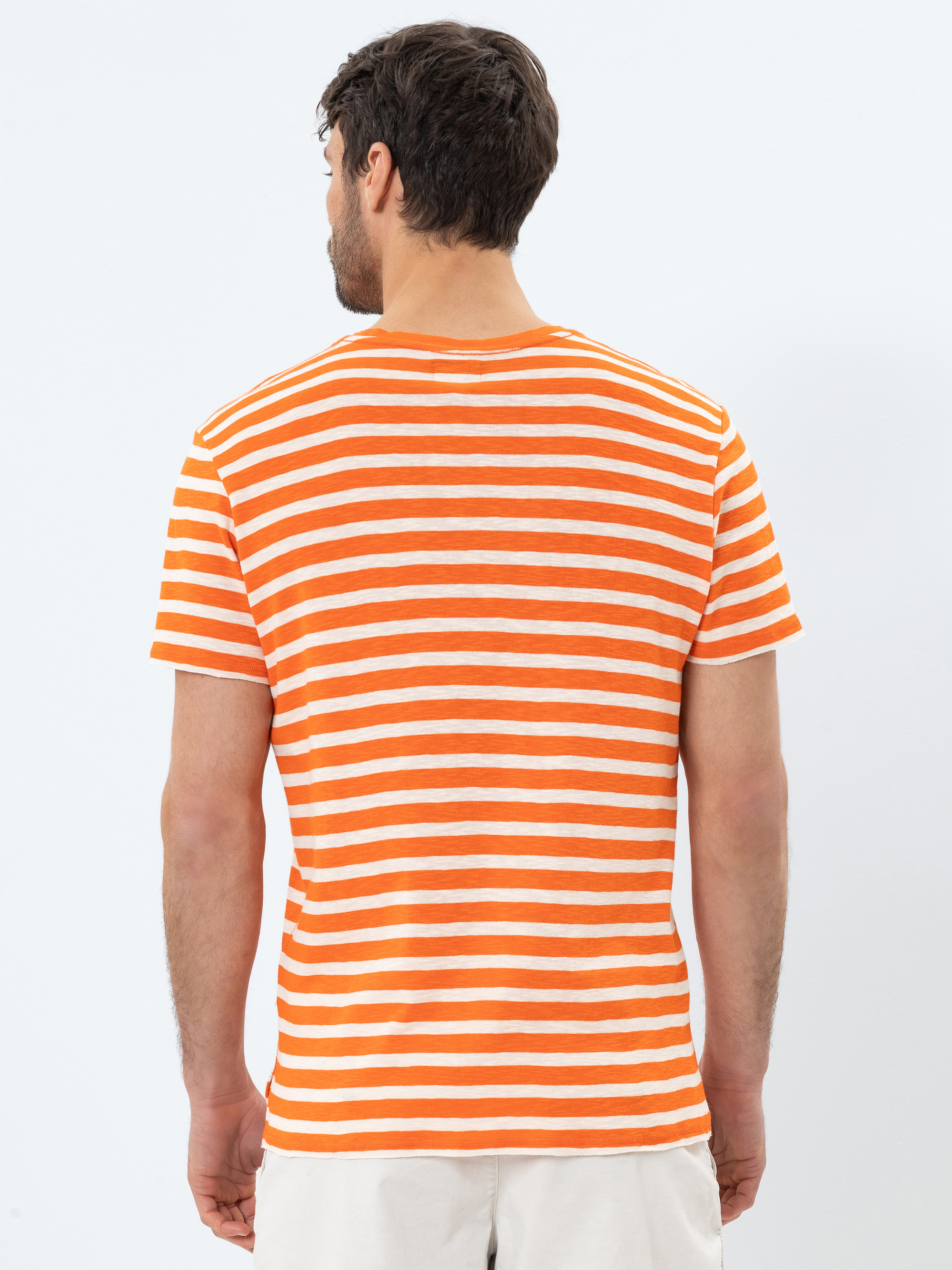 Ein Mann steht mit dem Rücken zur Kamera, trägt ein kurzärmeliges orange-weiß gestreiftes T-Shirt und helle Shorts vor einem schlichten weißen Hintergrund.