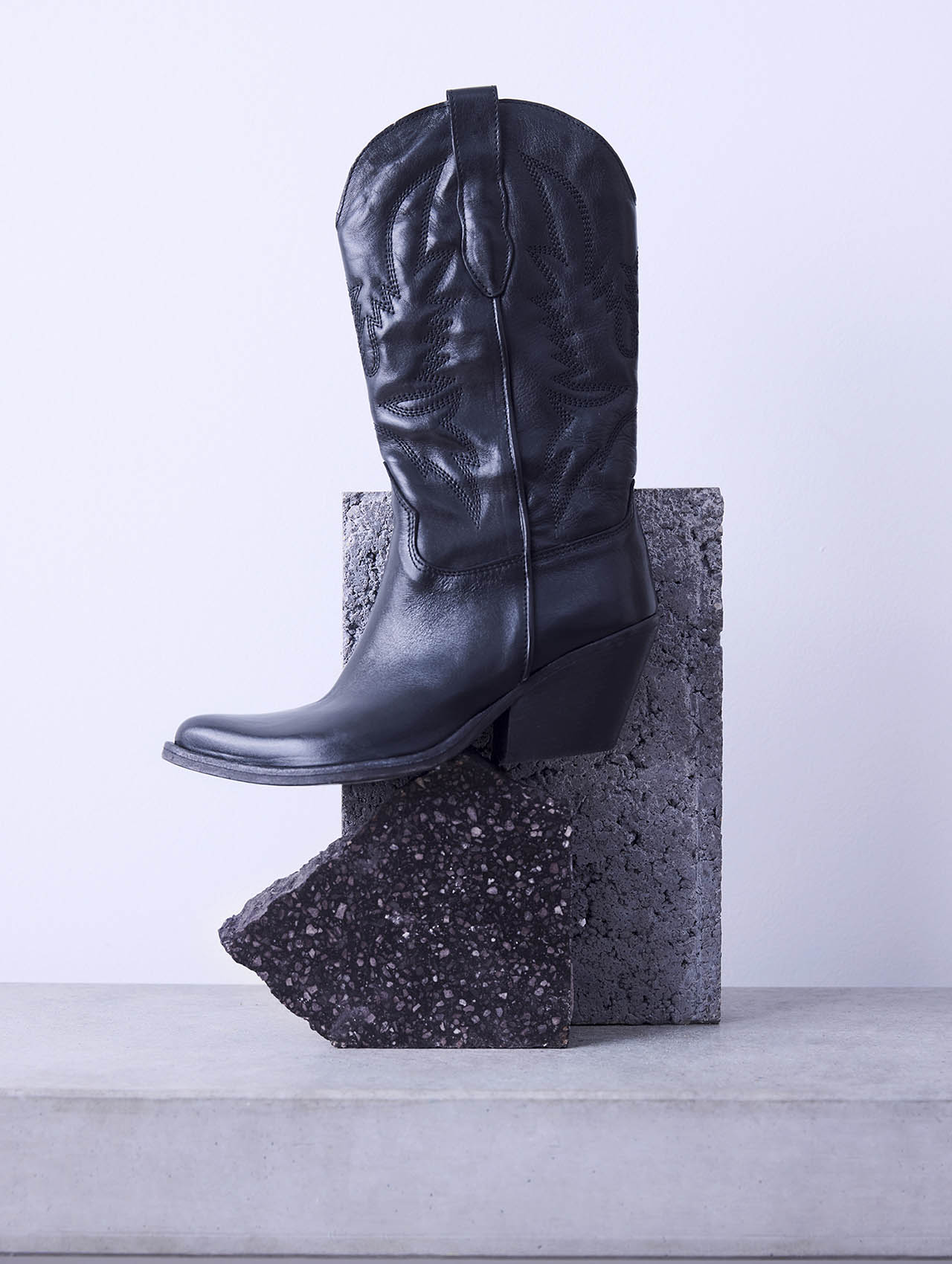Ein schwarzer Cowboystiefel aus Leder mit Ziernähten, der aufrecht auf geometrischen Steinblöcken vor einem hellgrauen Hintergrund steht.